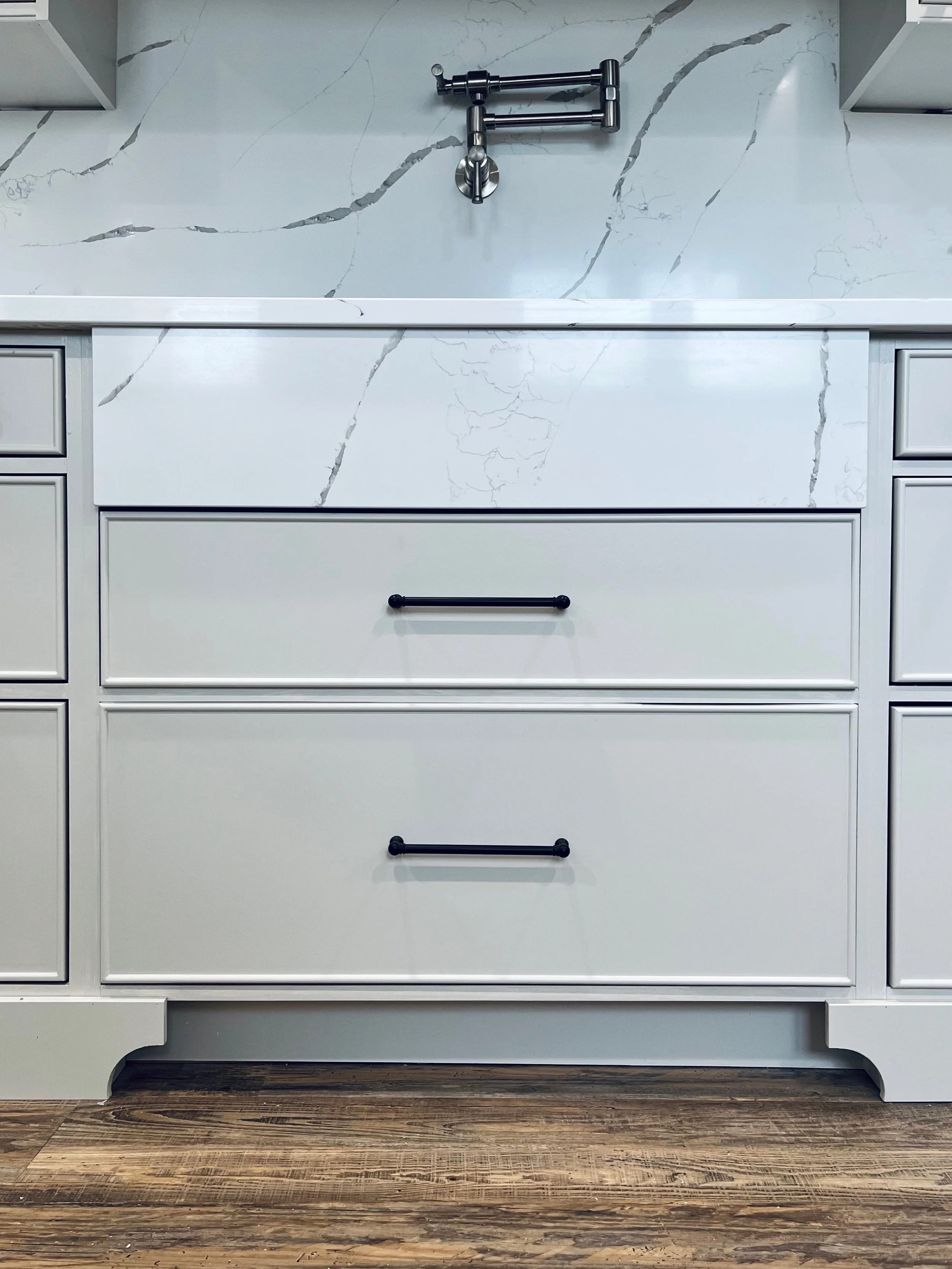 White kitchen vanity with two large drawers featuring black handles, a white marble countertop, and a marble backsplash with gray veining. A metallic faucet is mounted on the wall above the sink area.