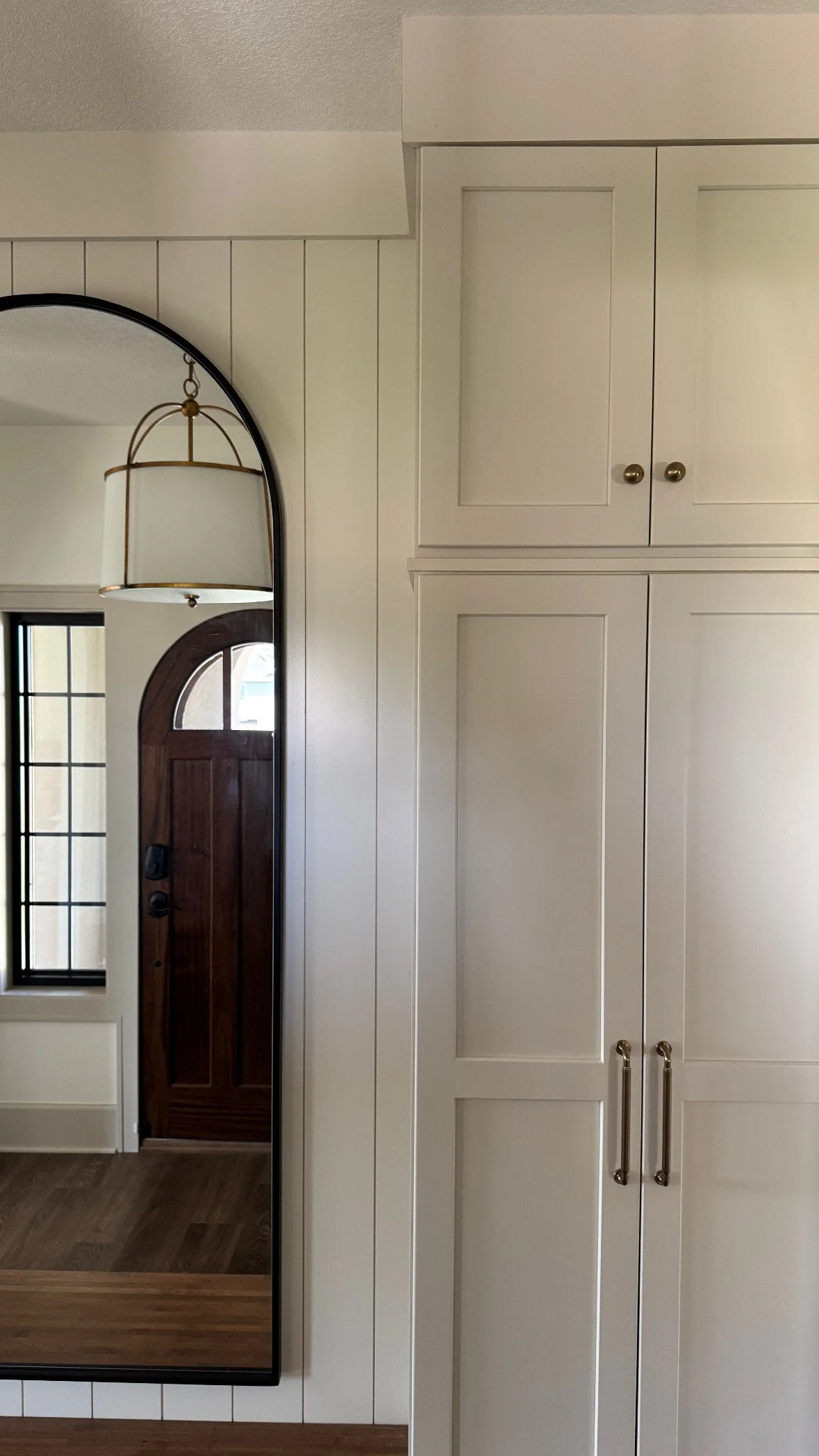 Interior view of a room with white cabinets, a large mirror, and a wooden front door reflected in the mirror.