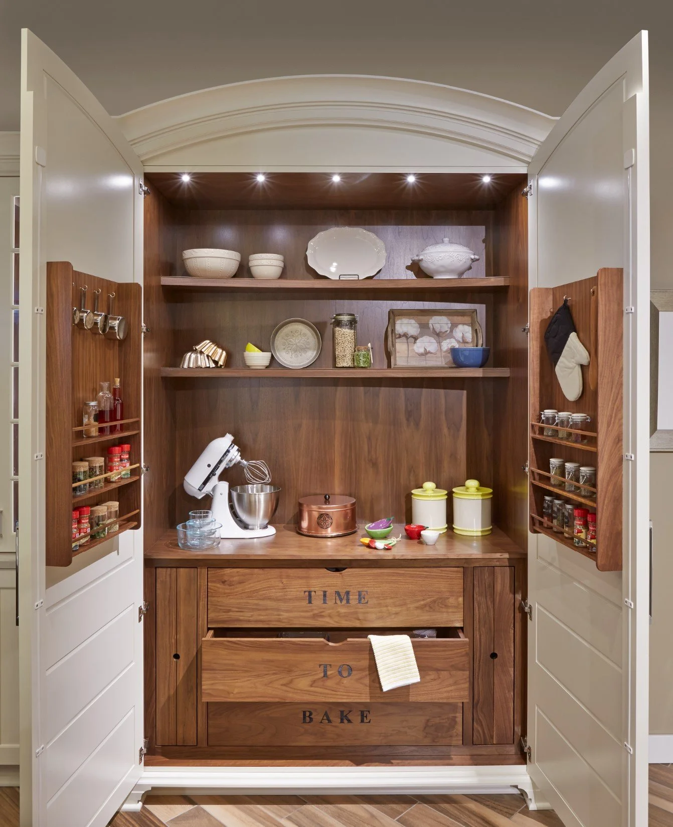 A wooden kitchen cabinet with open doors, containing shelves with bowls, jars, and decorative items, plus a countertop with kitchen appliances, dishes, and baking supplies.