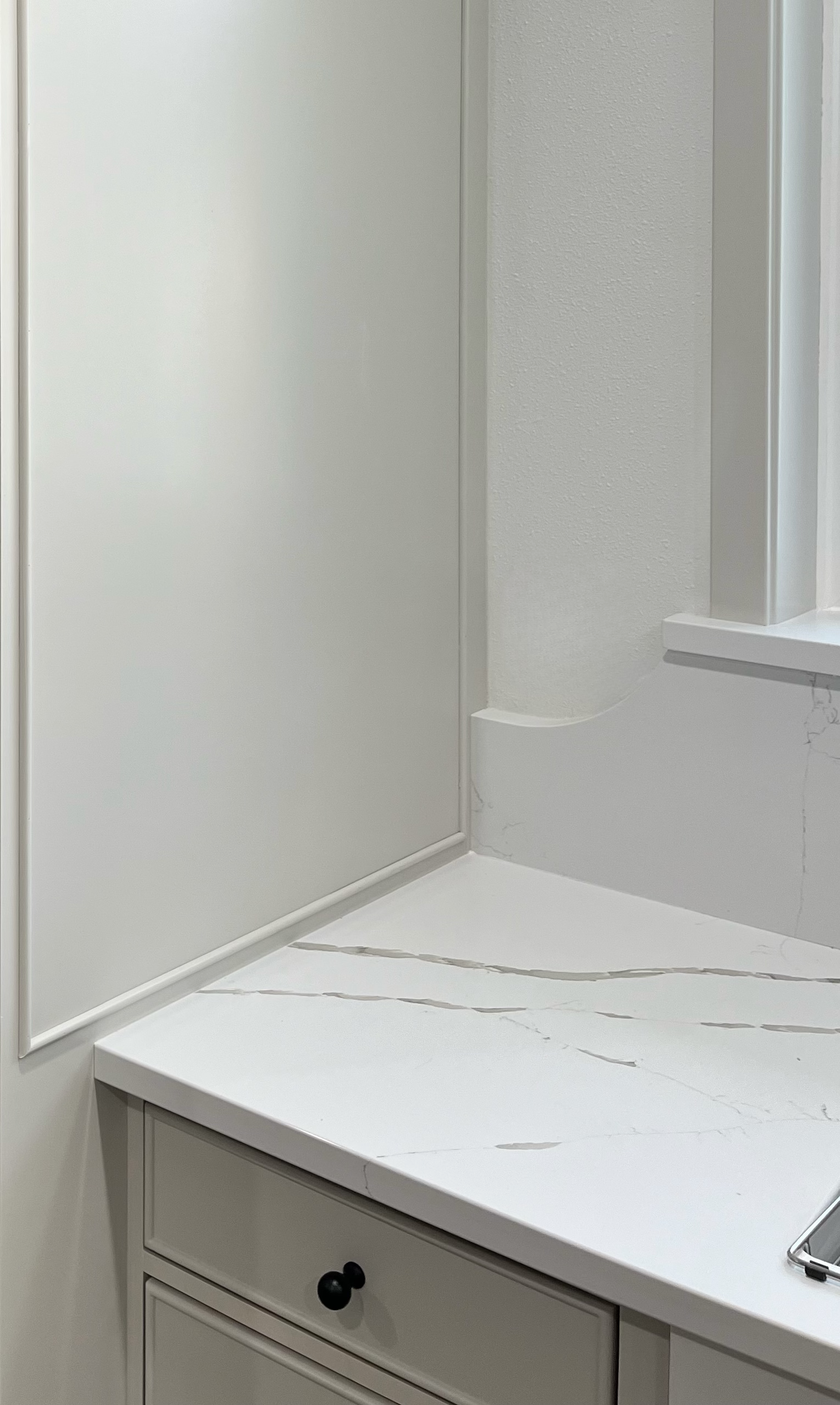 Close-up of a white bathroom countertop with a marble-like pattern, a cabinet with a black knob, and part of a mirror on the wall.