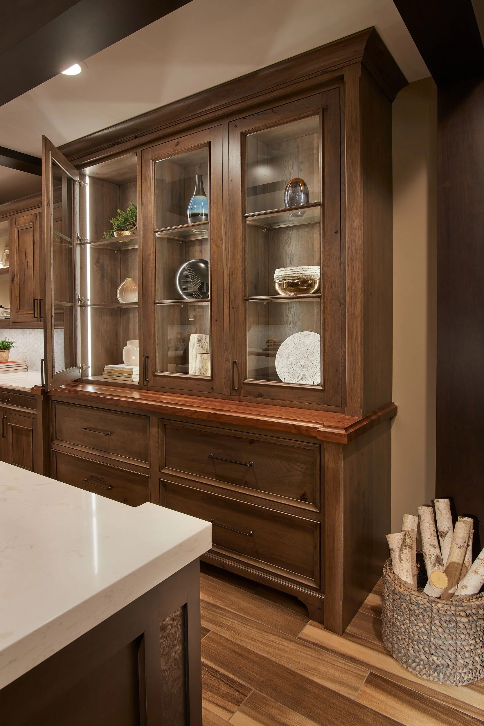 A wooden kitchen cabinet with glass doors displaying decorative vases and dishes, located in a home kitchen with wood flooring and a basket of firewood nearby.