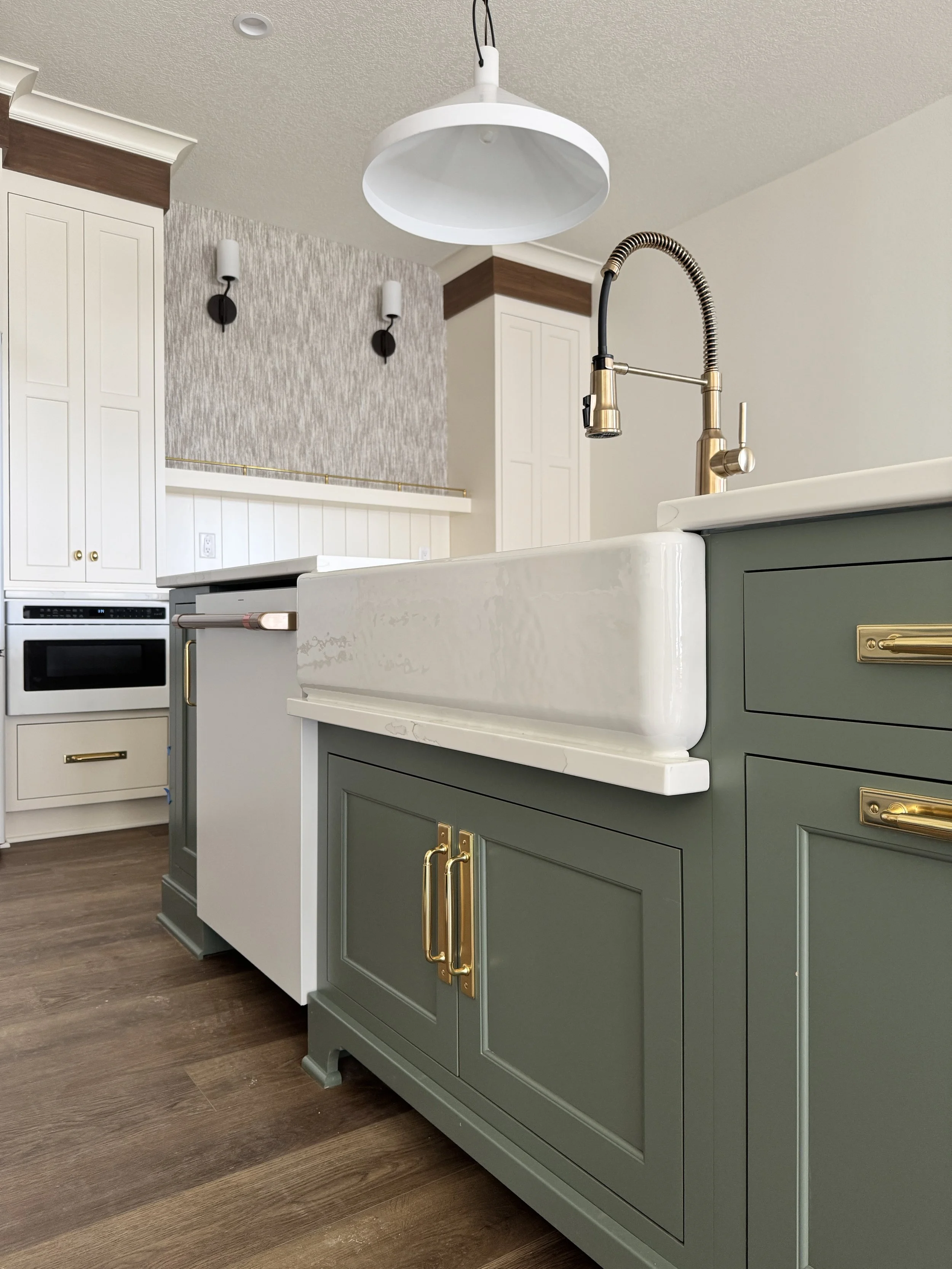 Close-up of a modern kitchen sink with a rustic farmhouse style, featuring a cream-colored apron sink, green cabinetry with gold hardware, and a gooseneck faucet, with a white pendant light overhead and a wood floor.
