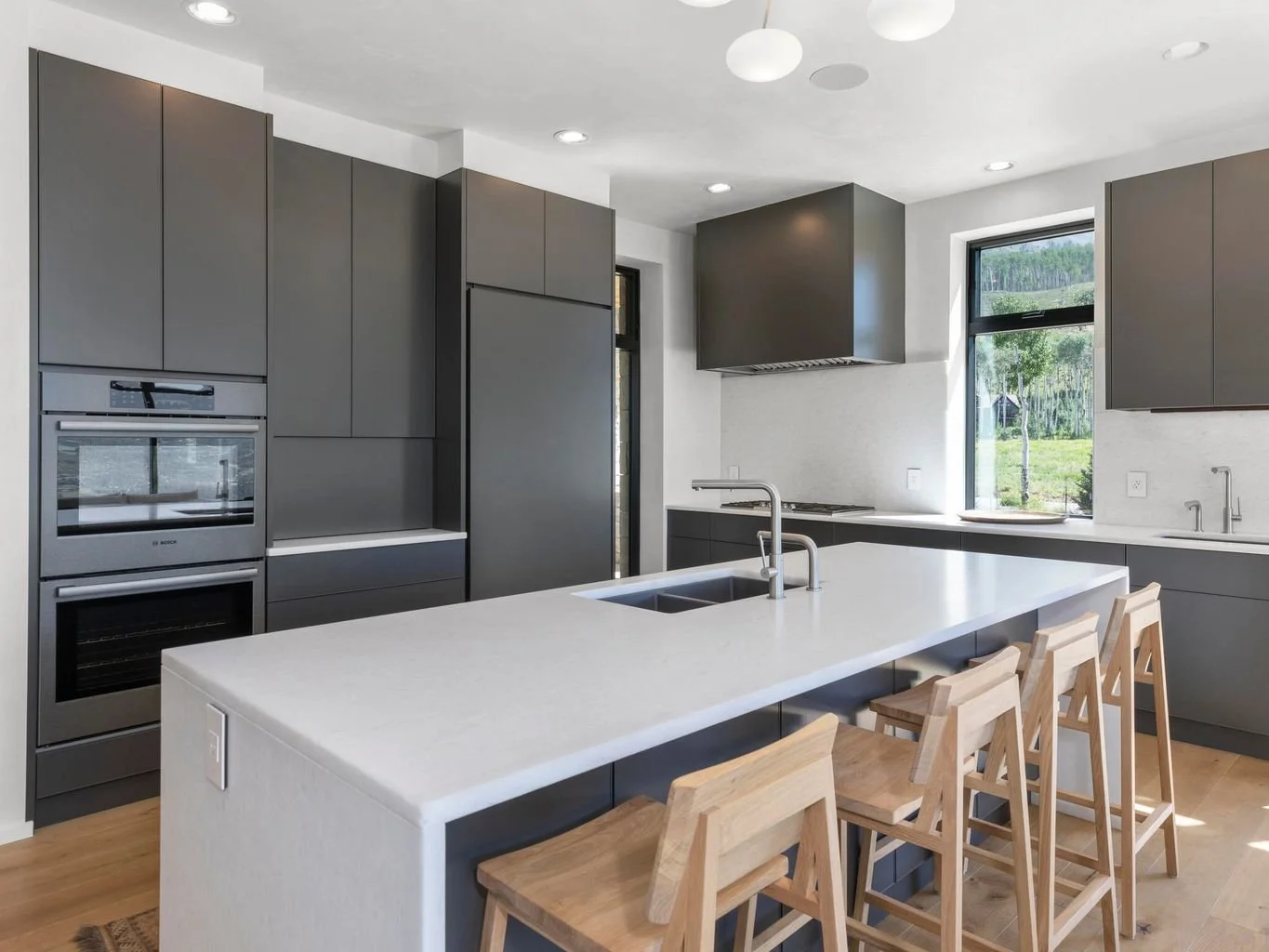 Modern kitchen with gray cabinets, white countertop island, wooden chairs, stainless steel appliances, and large windows showing outdoor greenery.