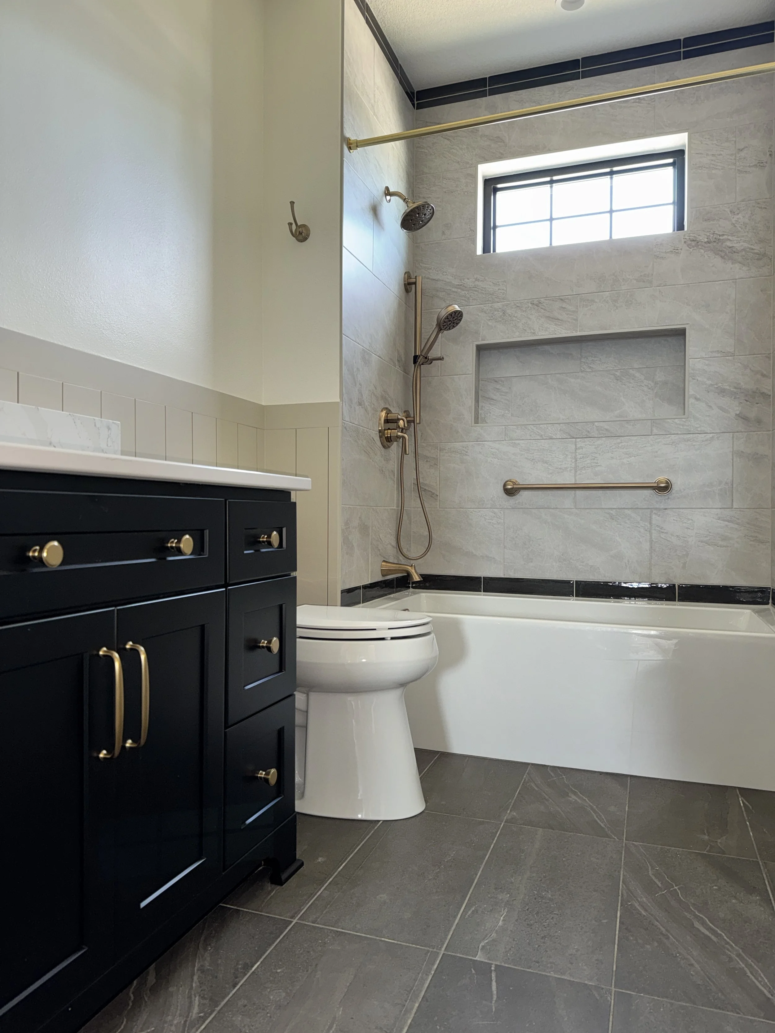 Modern bathroom with a dark blue vanity, white countertop, and a toilet next to a bathtub. The bathtub area has a shower with corn gold fixtures, a niche, and a window above.