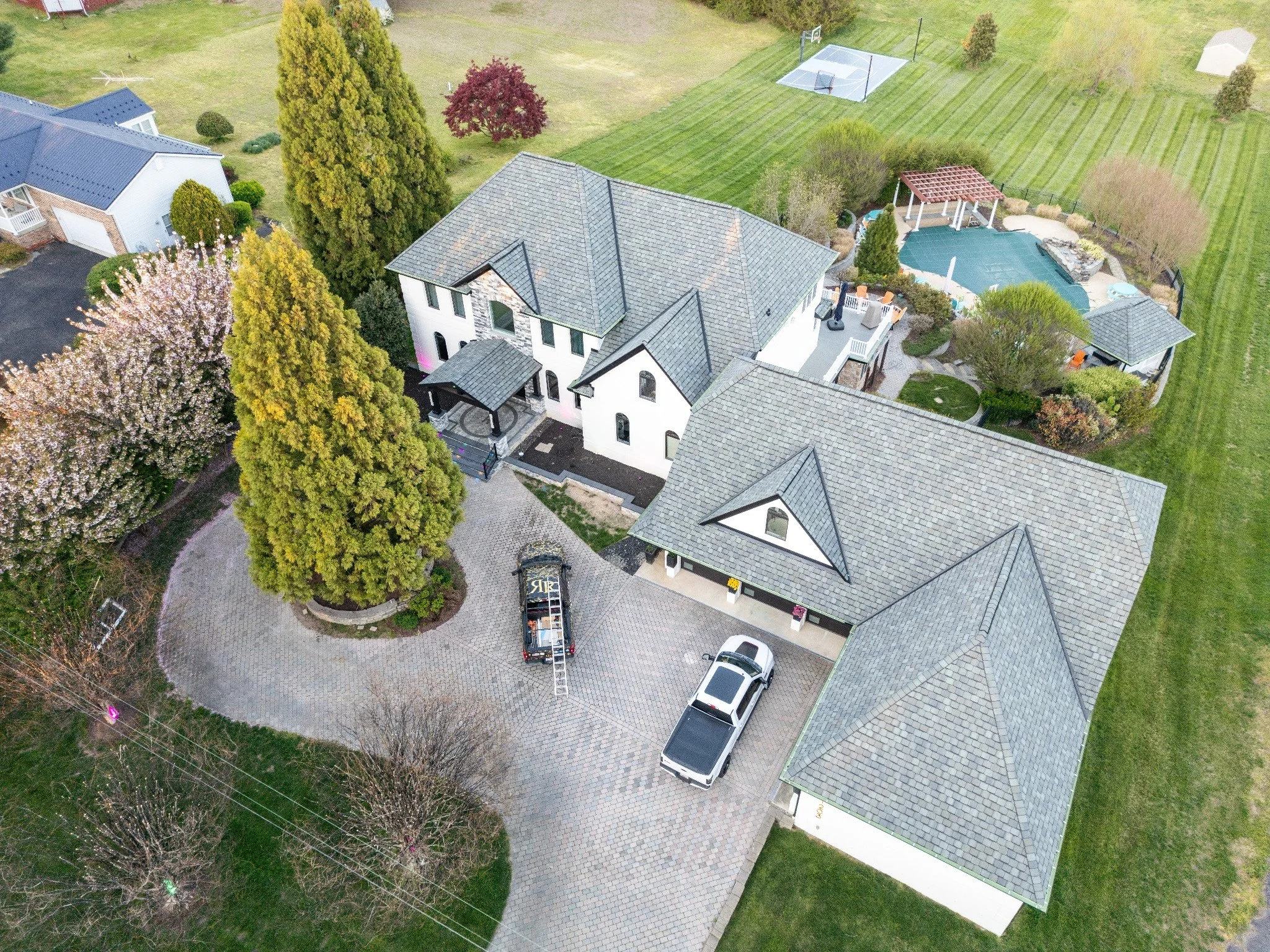 Aerial view of a large house with multiple gray gabled roofs, surrounded by a driveway, trees, and a backyard with a tennis court and a swimming pool.
