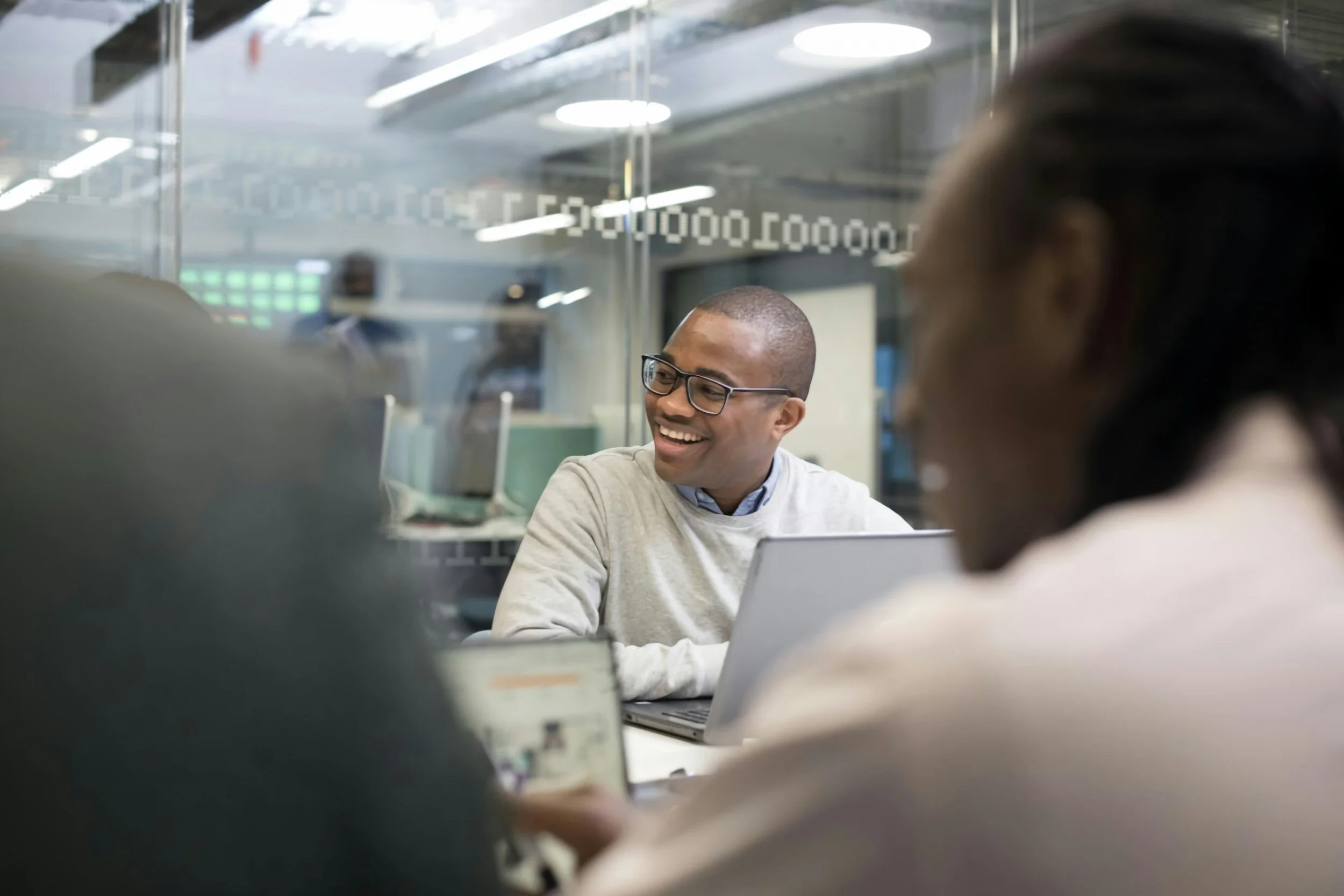 People in a modern office having a meeting, one person smiling and wearing glasses, with laptops on the table.