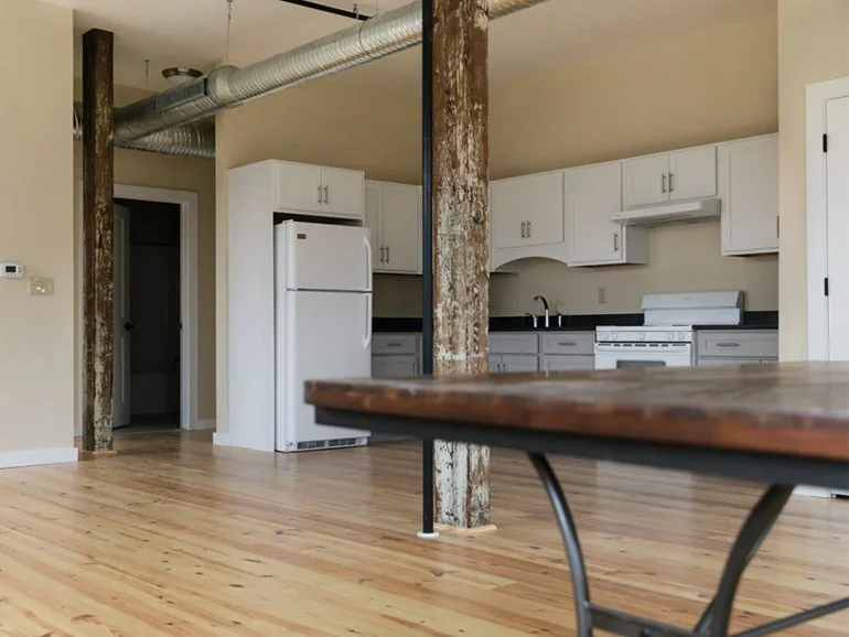 Loft apartment interior with exposed beams, white kitchen cabinets, refrigerator, stove, and wooden flooring.