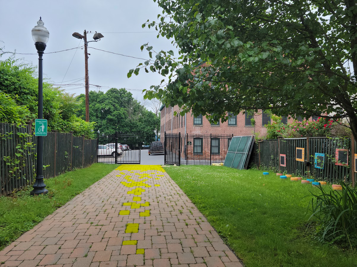 Brick pathway with yellow markings, leading to a gated parking area, surrounded by green grass, fences, and trees, with power lines overhead.