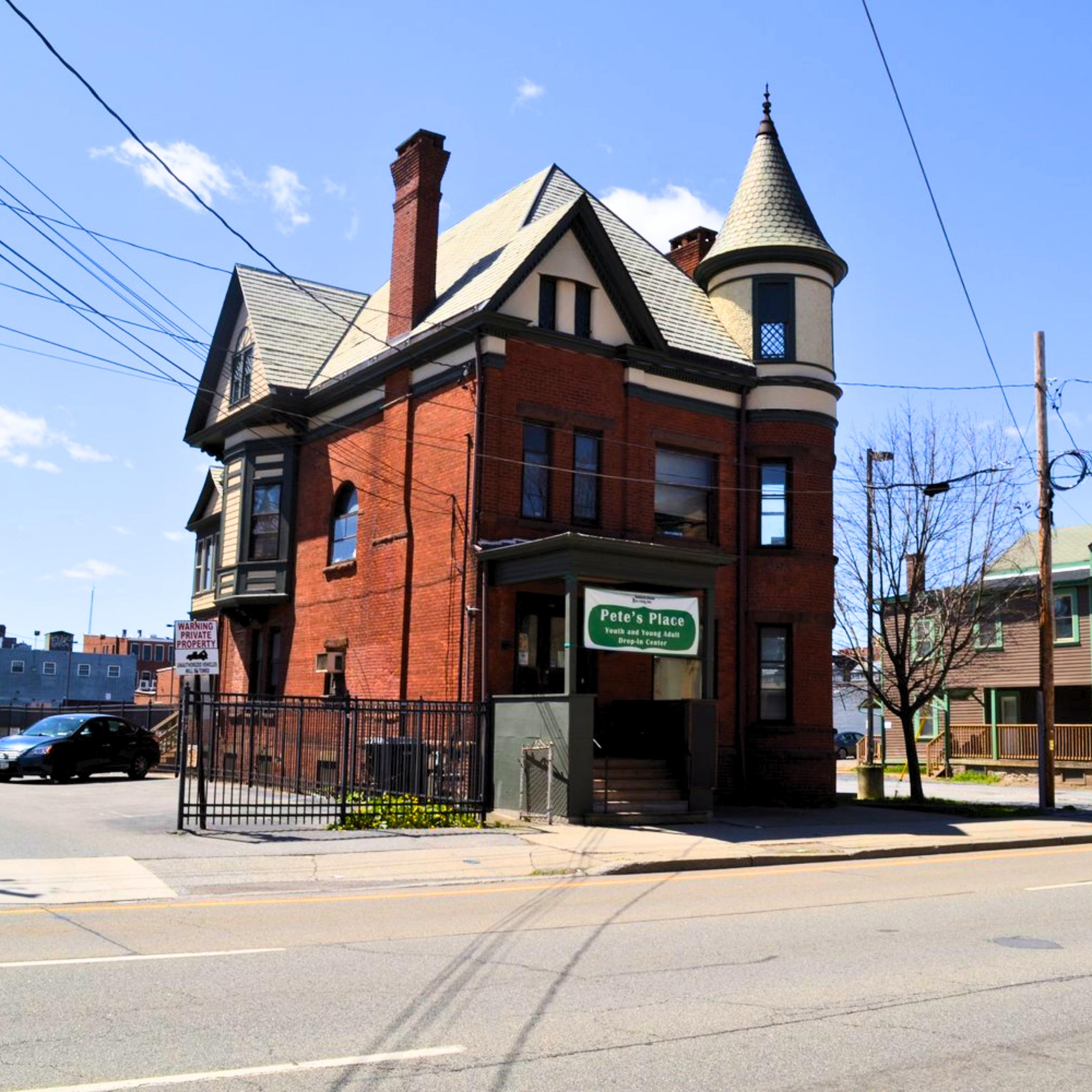 Street view of a modern building with large windows and a wooden door, featuring industrial-style lamps and a red fire hydrant nearby. The sidewalk is wet, indicating recent rain, and there's a tree and other buildings in the background.