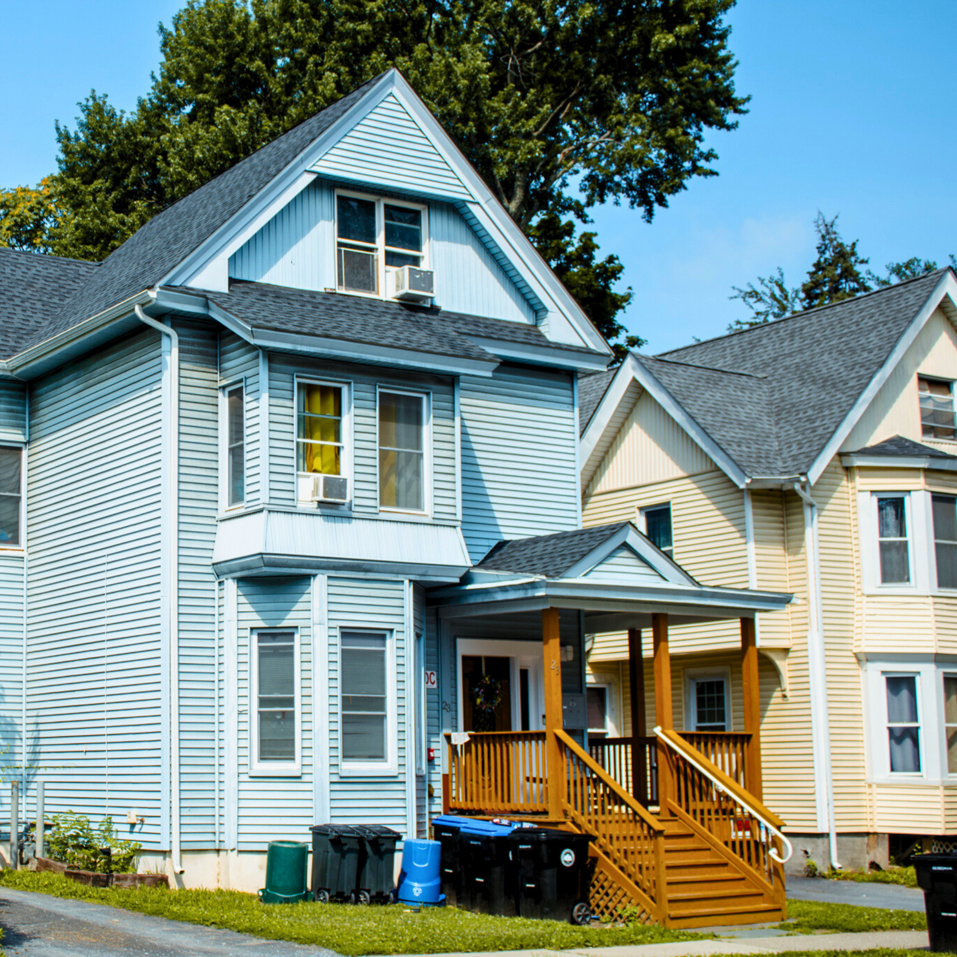 Victorian-style house with a porch, multiple windows, and mansard roof, surrounded by greenery and a clear blue sky.