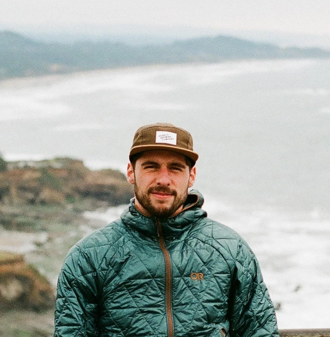 A man wearing a green quilted jacket and a brown cap standing on a coastal cliff with waves crashing in the background.