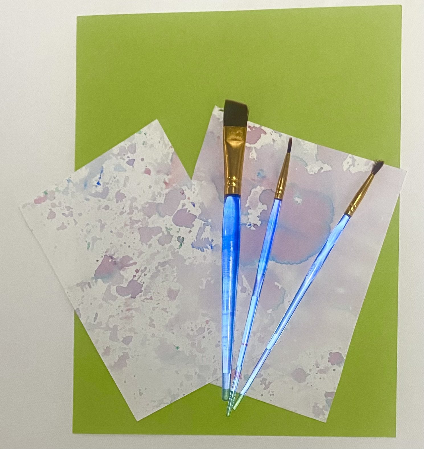 Three paintbrushes with glowing blue handles resting on watercolor paper on a green background.