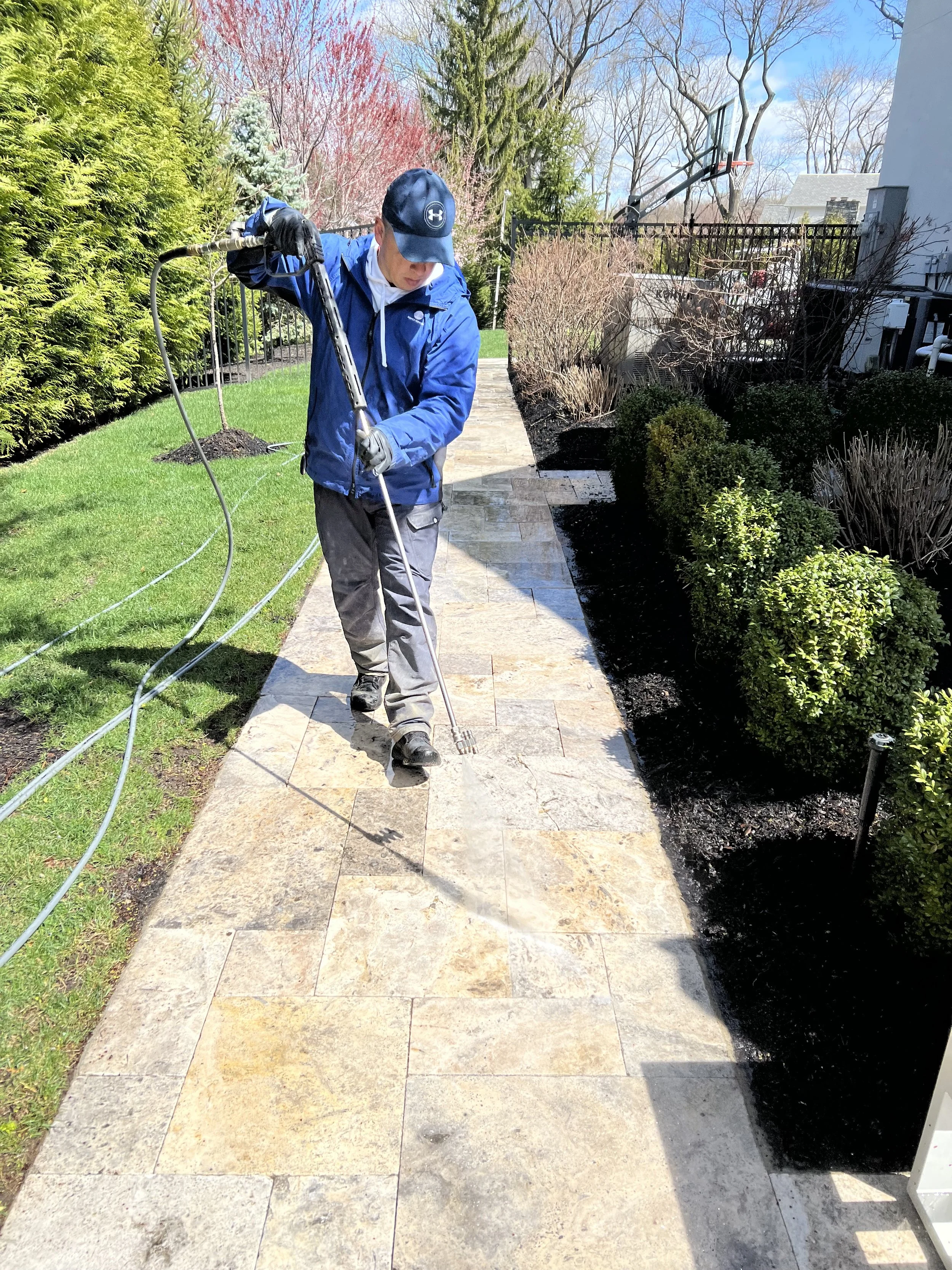 A person in a blue jacket, black cap, and gloves power washing a stone pathway in a garden on a sunny day, with landscaped bushes and trees in the background.