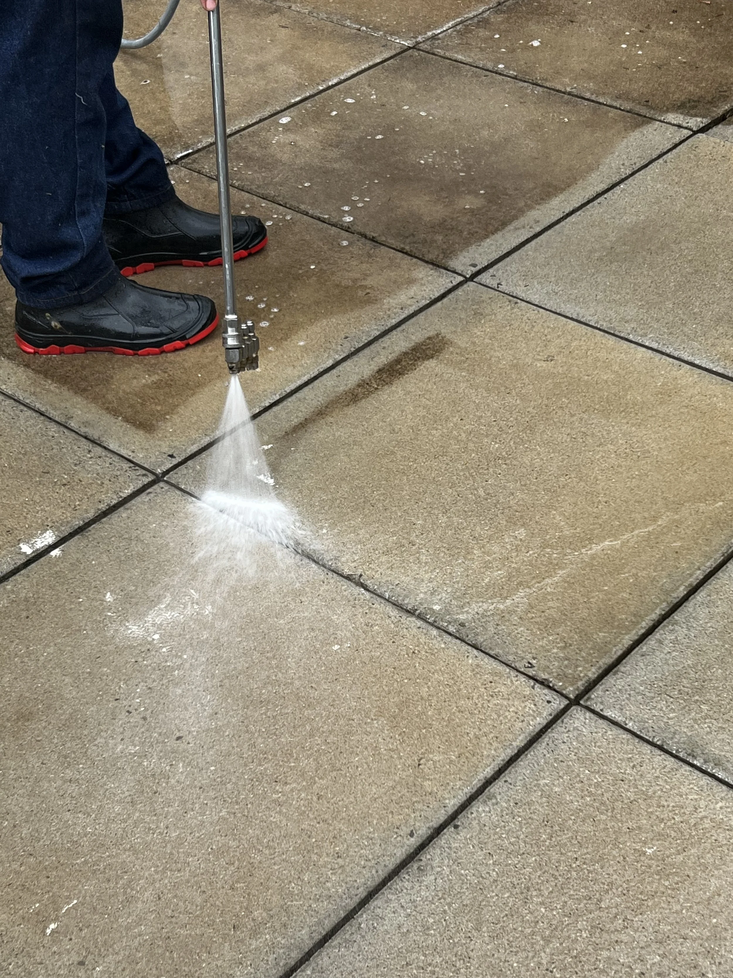 A person wearing black shoes with red soles is using a pressure washer to clean a concrete sidewalk.