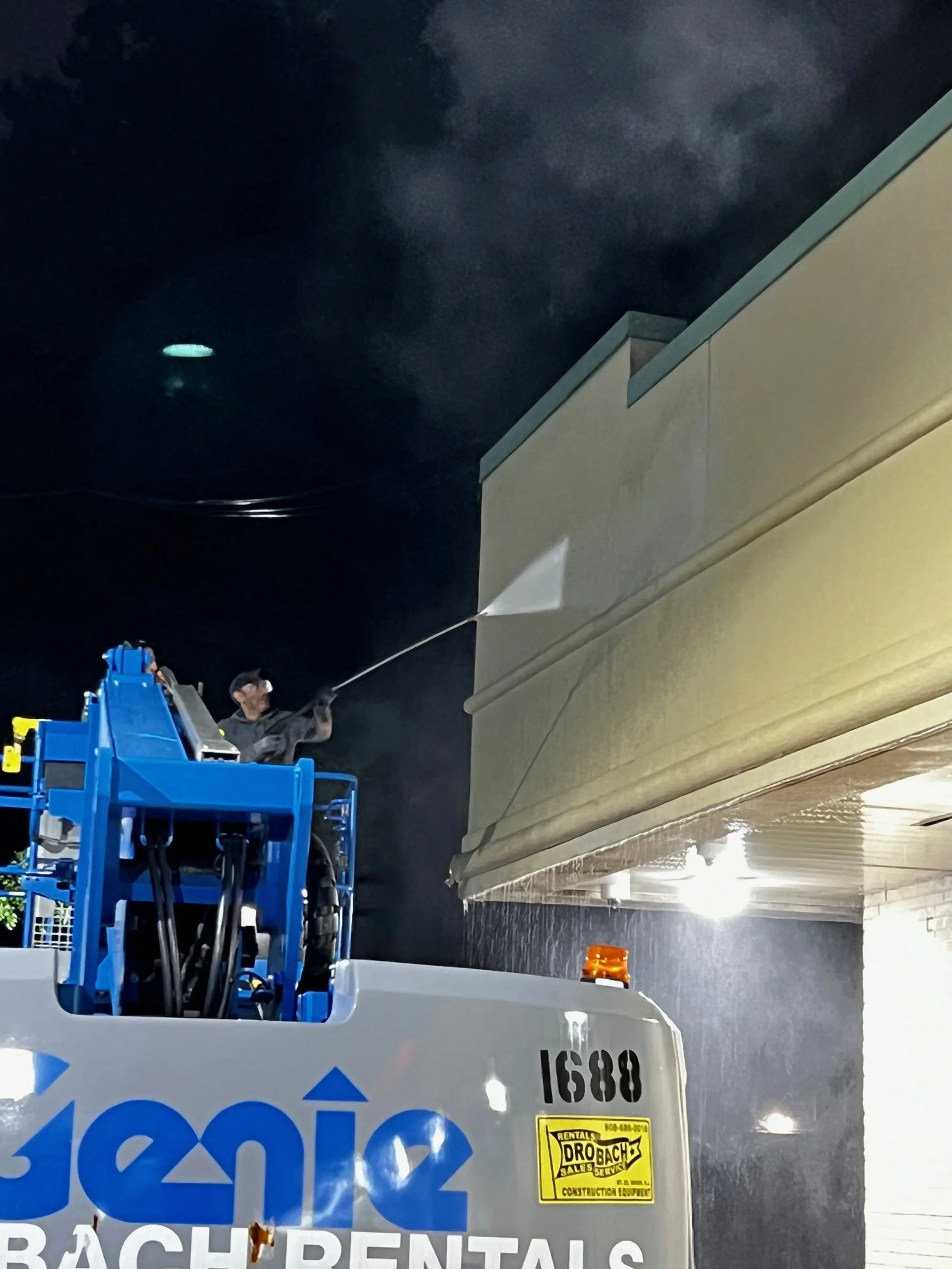 A worker cleaning the exterior of a building with a pressure washer at night. The worker is standing on a blue lift platform.