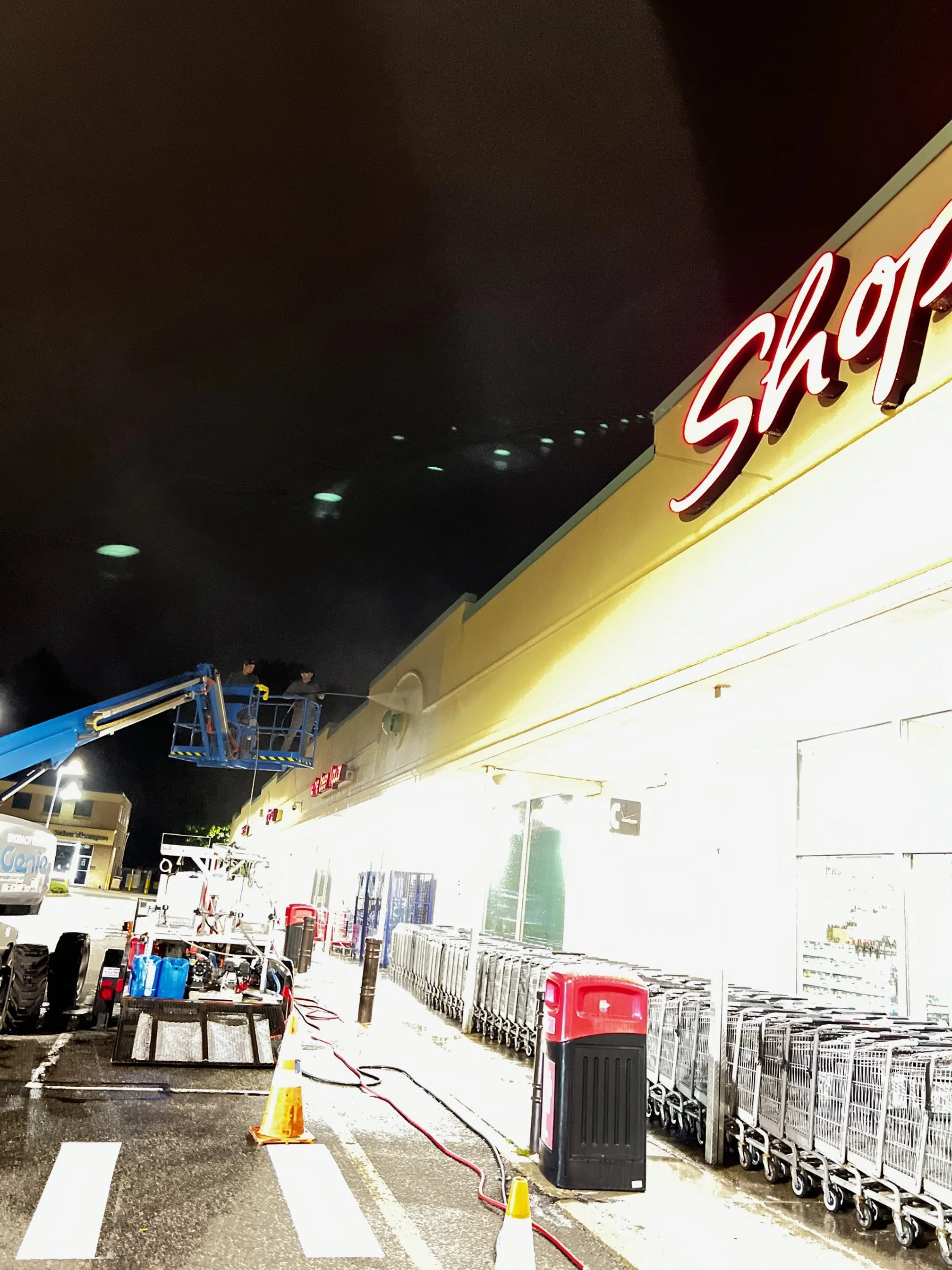 Nighttime scene outside a supermarket with a bright, illuminated storefront and shopping carts lined up. Maintenance work is underway with workers on a lift, and cones and equipment are set up on the parking lot.