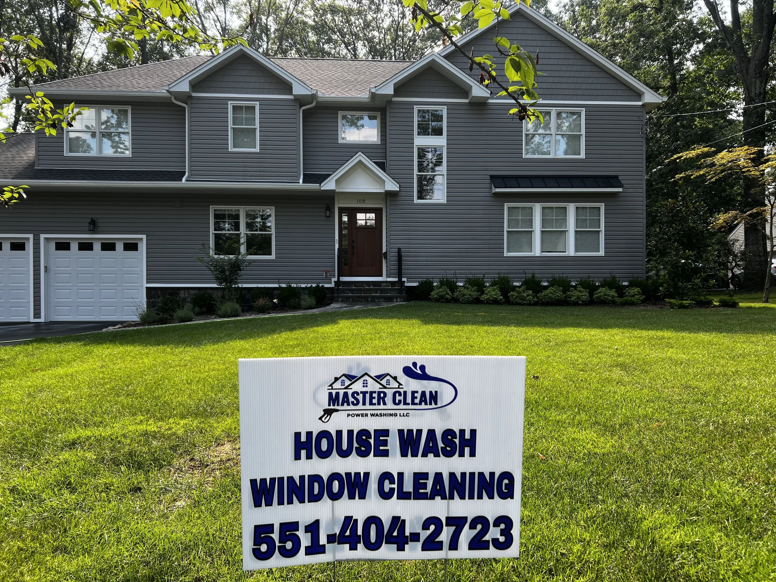 A two-story gray house with white trim and a brown front door, surrounded by a green lawn, with a sign in the foreground advertising house washing and window cleaning services.