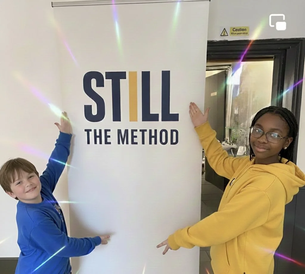 Two children, a boy in a blue jacket and a girl in a yellow hoodie, are standing next to a white sign that says "STILL THE METHOD." The children are smiling and pointing towards the sign. There are colorful light effects in the image.