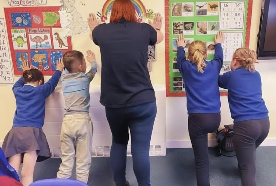Four children and one teacher in a classroom, all placing their hands on a wall with educational posters about animals and a world map.