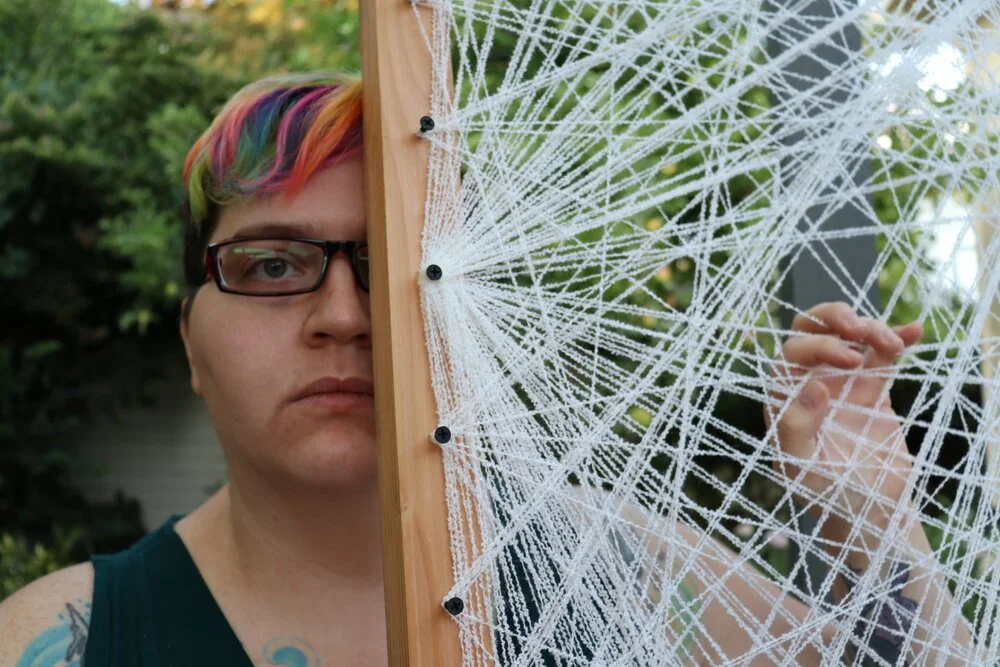 Rae Maxwell-Ross, a non-binary person with show rainbow dyed hair and rectangular glasses, holds a large spiderweb-like artwork over the left side of their face.