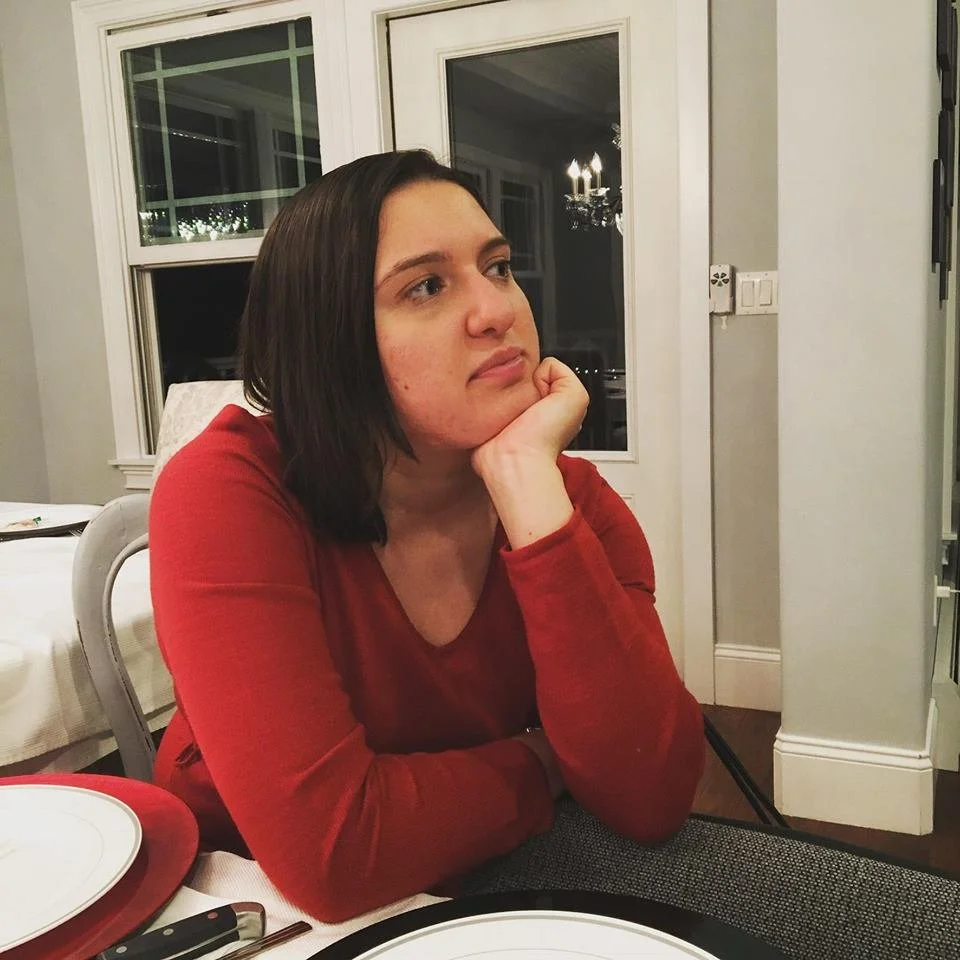 Amanda DeLalla sitting in at table. She wears a red shirt and rests her chin in the palm of her hand