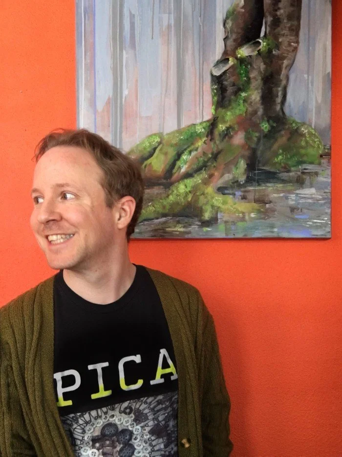 Josh Gates posing with one of his painting, smiles and looks off to the side