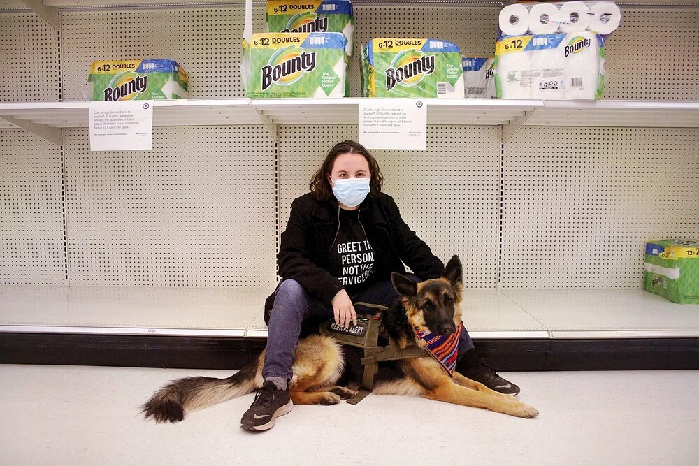 Oaklee Thiele sits at the bottom of an nearly empty store shelf with her service dog, Coco.