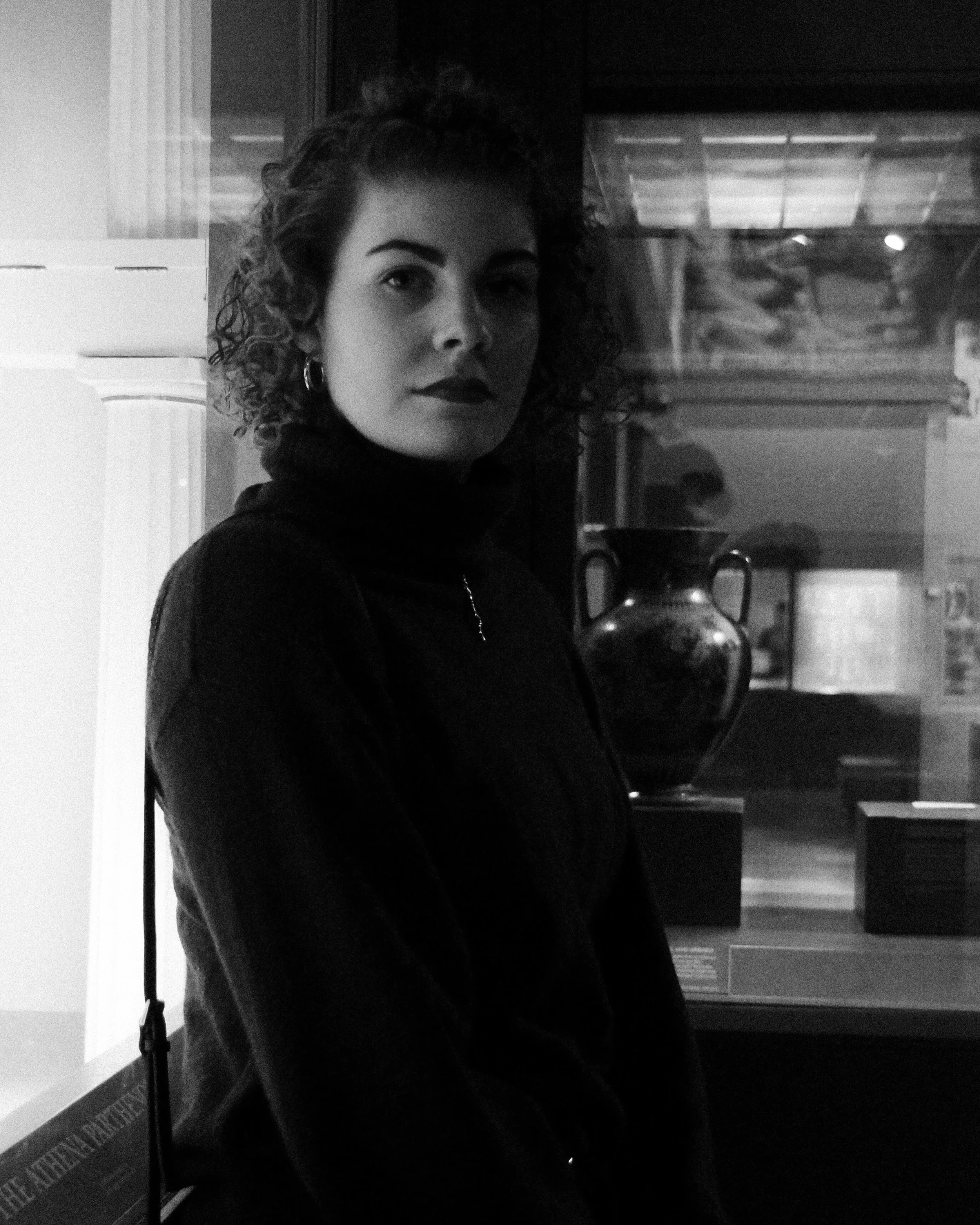A black and white image of a white woman with dark curly hair wearing a turtleneck sweater and leaning back against a glass museum display case.