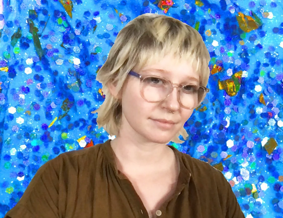A blonde, pale, non-binary person smiles at the viewer.