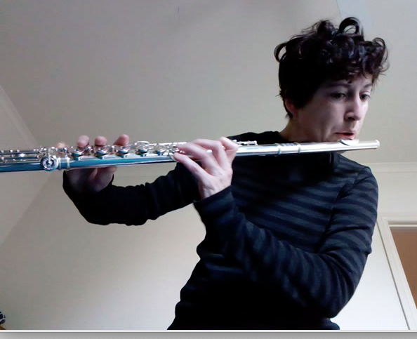 Mel, a gender nonconforming woman with short dark curly hair plays a flute.