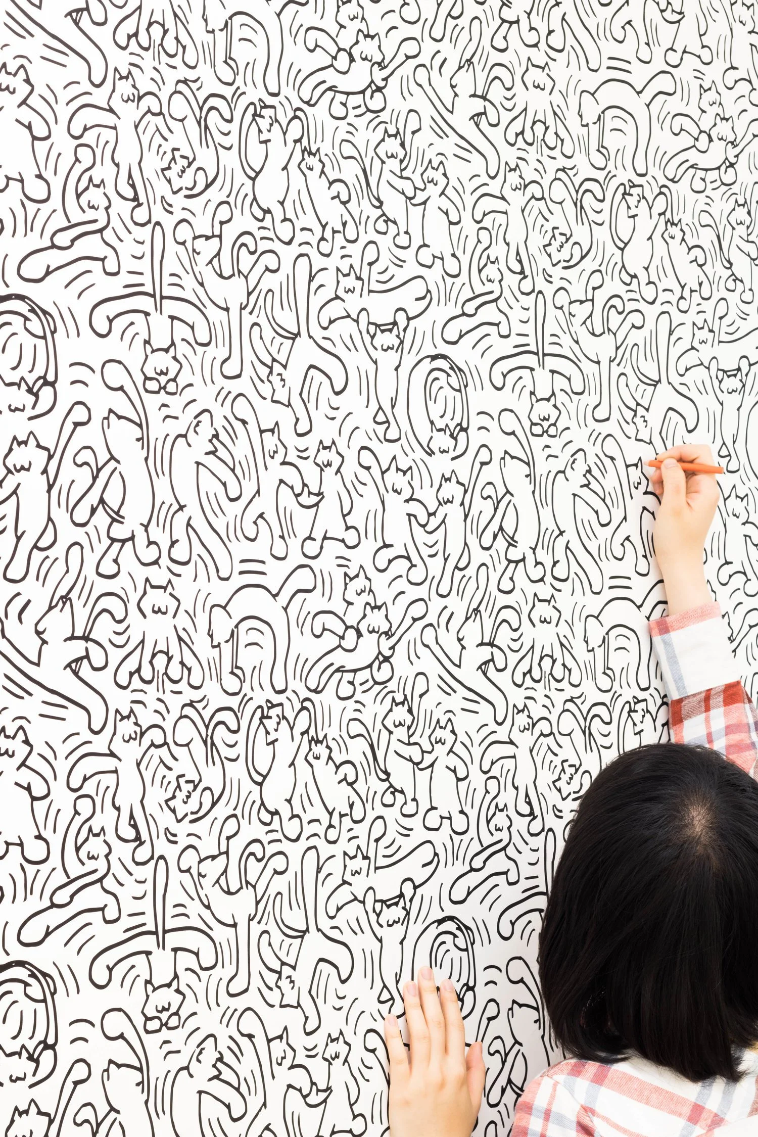 The artist, seen from behind, reaching up and drawing multitudes of dancing cats on a white wall.