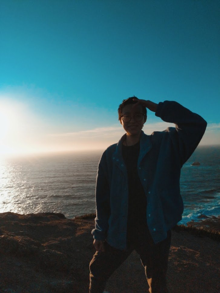 A Việt-American person posing with one hand scratching their forehead. Behind them is a sunset and the ocean.