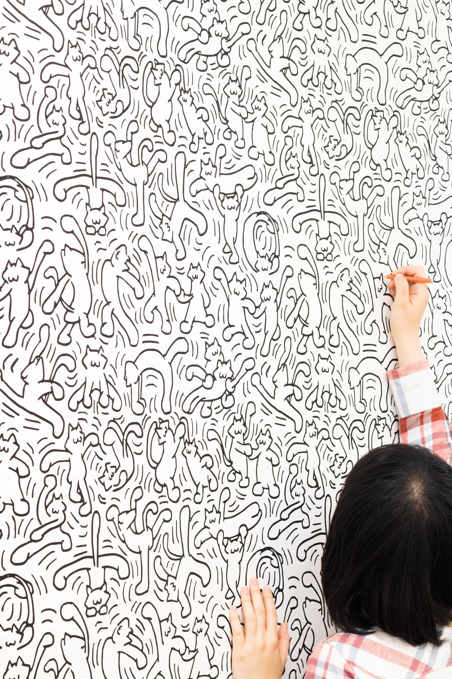 The artist, seen from behind, reaching up and drawing multitudes of dancing cats on a white wall.