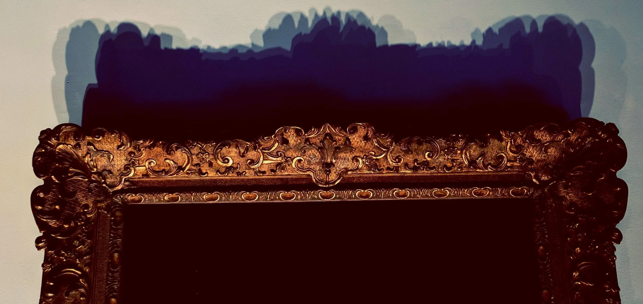 A close-up of a clean white gallery wall. Cropped awkwardly in the bottom of the grainy photo, a gold, ornate antique frame casts overlapping shadows in blue, grey, and deep purple.