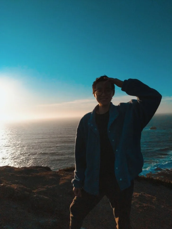 A Việt-American person posing with one hand scratching their forehead. Behind them is a sunset and the ocean.