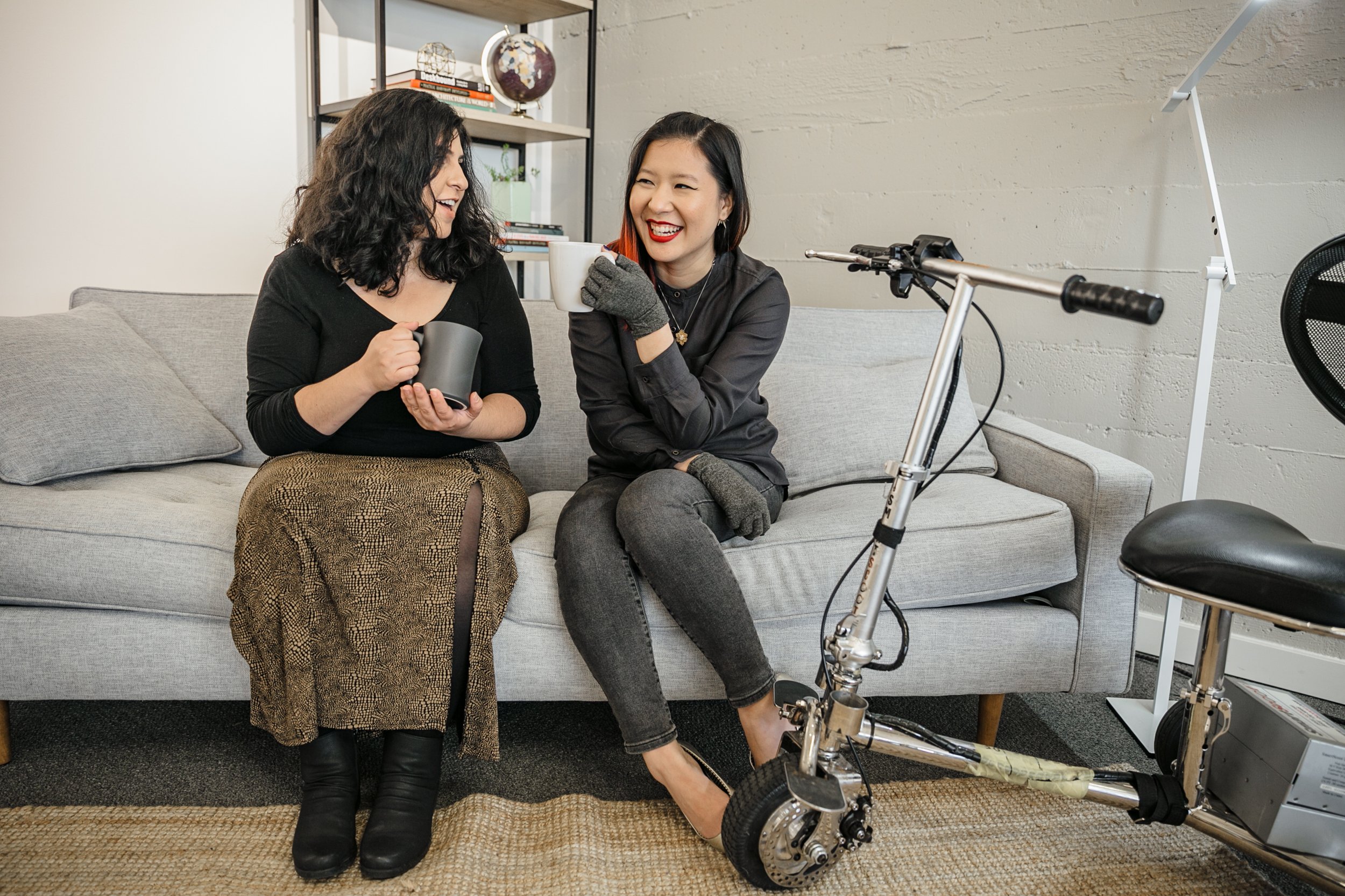 A Latinx disabled woman and an Asian disabled genderfluid person chat and sit on a couch.
