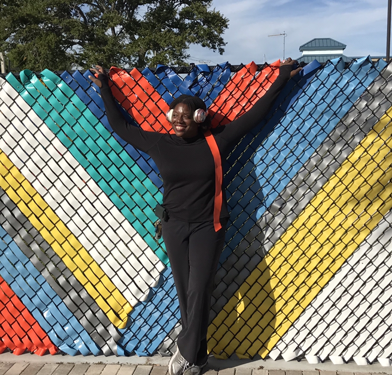 The Black Dalhia (Dalhia Perryman) stands in front of a colorful fence.