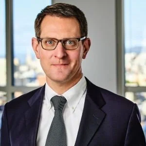A man with short brown hair, glasses, and a broad smile standing against a beige wall.