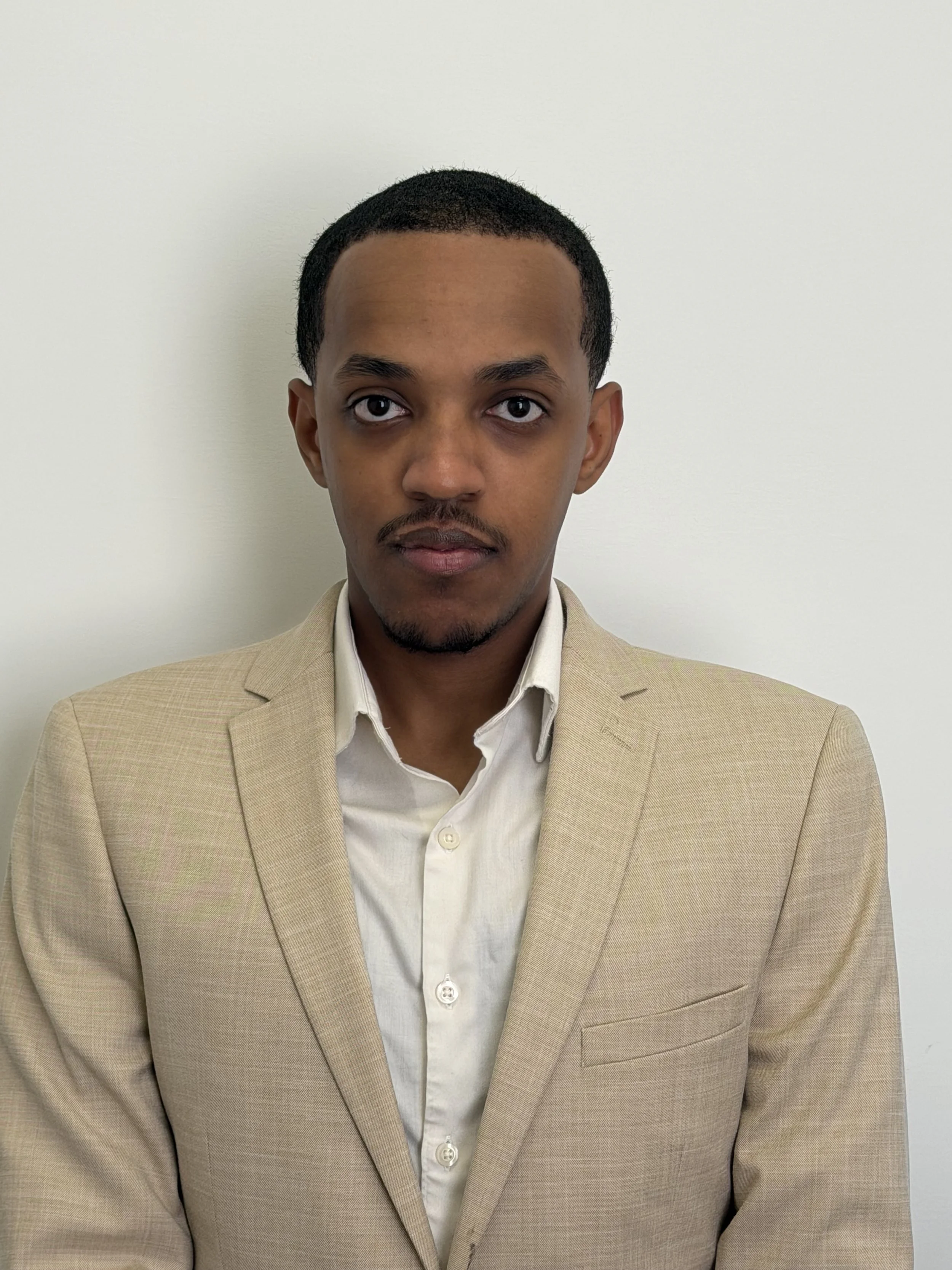 A person wearing a dark suit and a white shirt standing against a plain background.