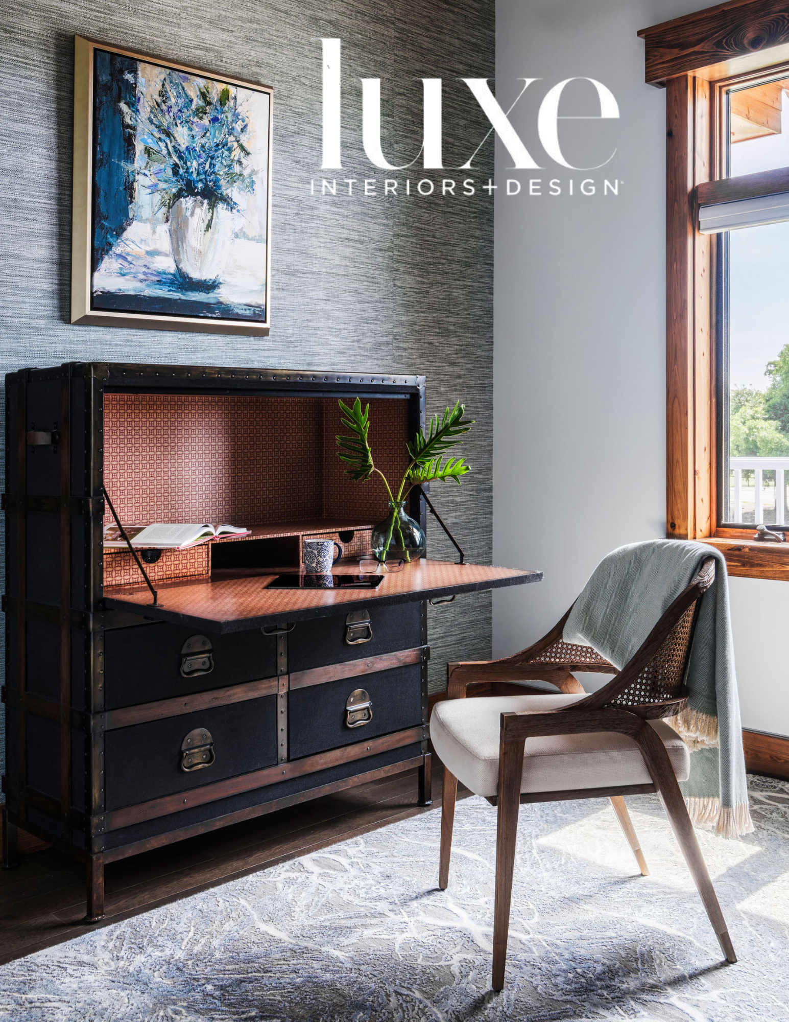 Interior of a room featuring a wooden chair with a cream cushion and a throw blanket, a large window with wooden trim, a blue and white patterned vase with green leaves on a black industrial-style credenza, a framed abstract painting of a flower in s
