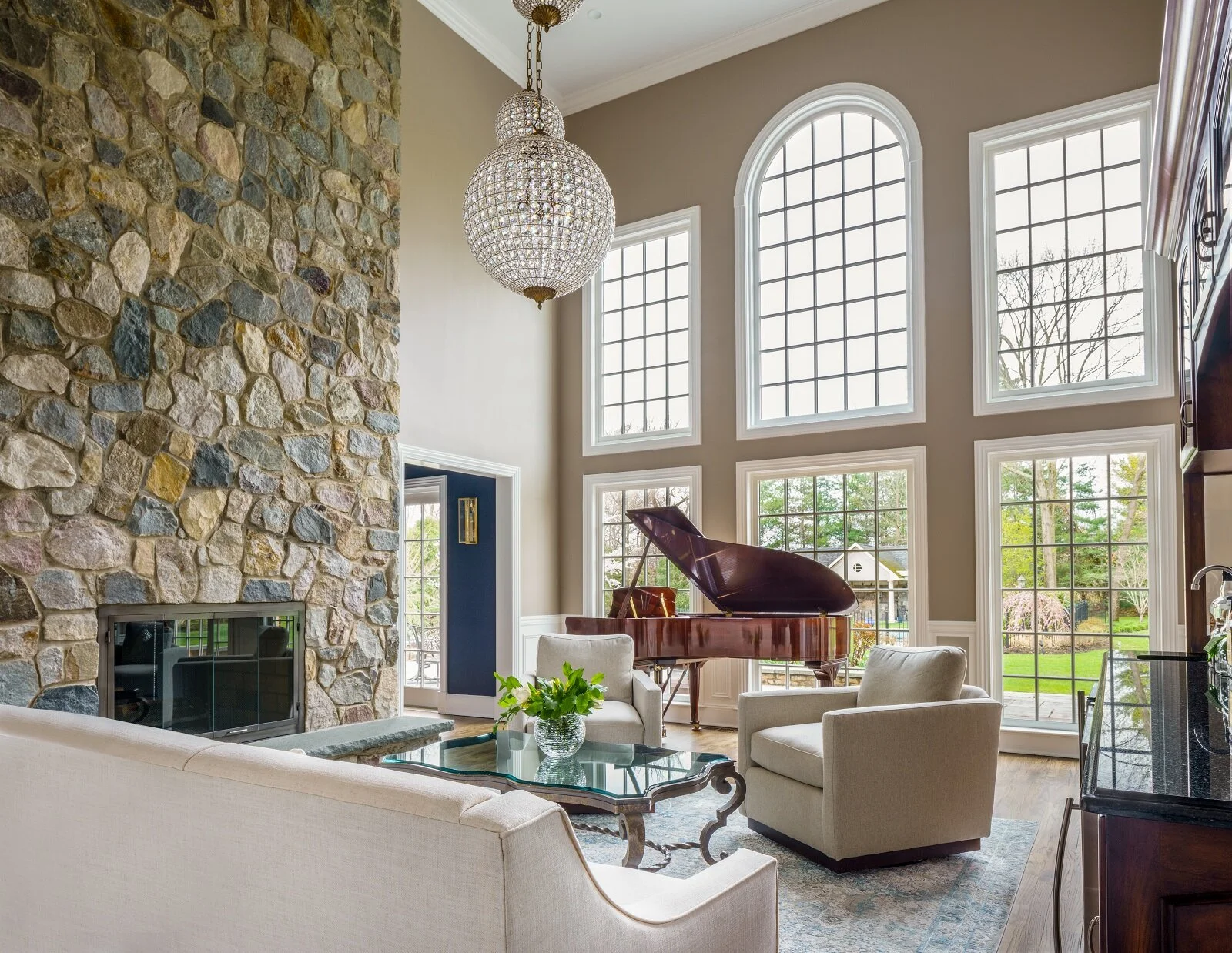 Living room view to french doors and windows. Stone fireplace, two chairs, cocktail table with flowers. Piano in back ground.