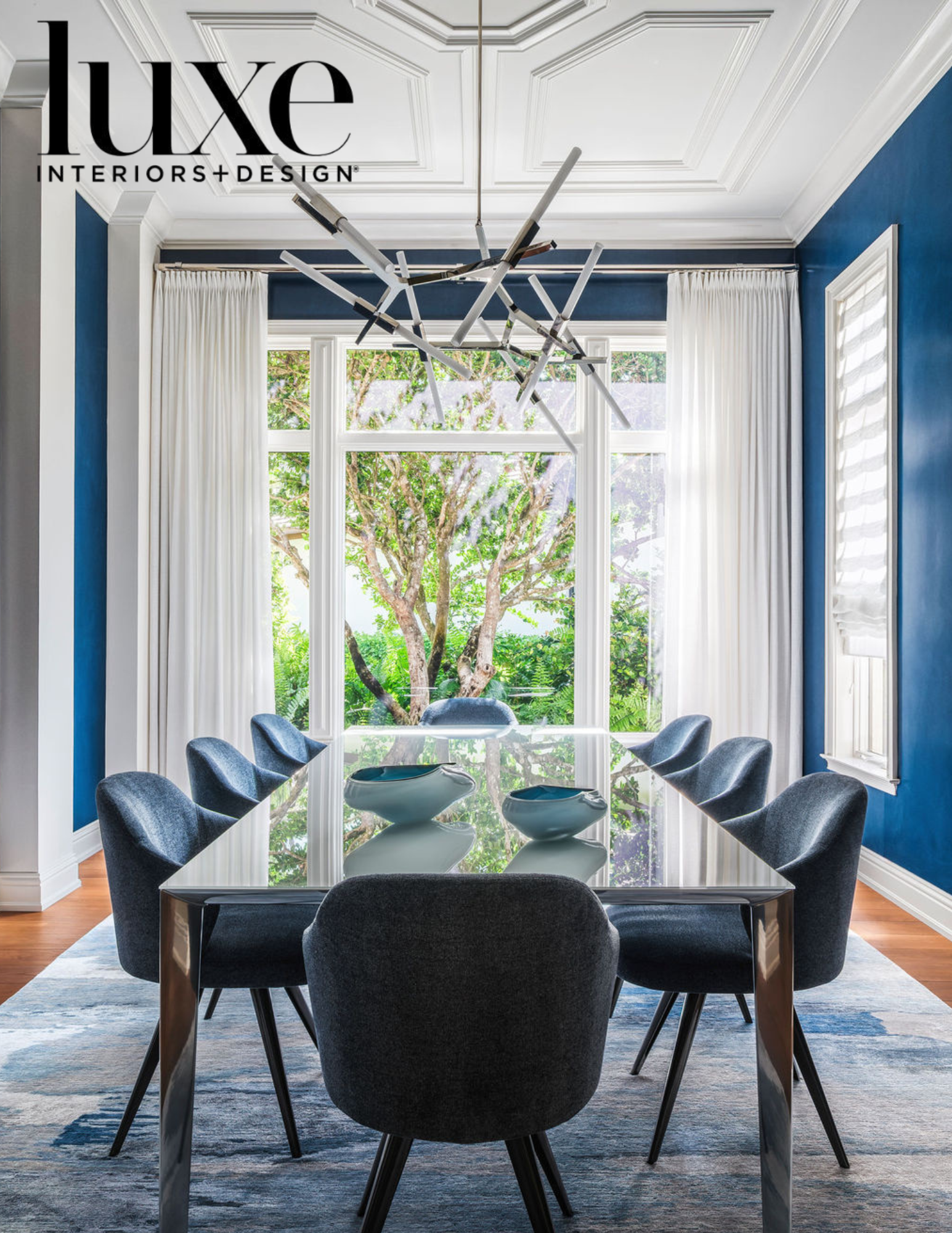 A modern dining room with a glass table, eight dark blue upholstered chairs, a large window with white curtains, a blue accent wall, a white ceiling with decorative molding, a contemporary black and white chandelier, and decorative bowls on the table