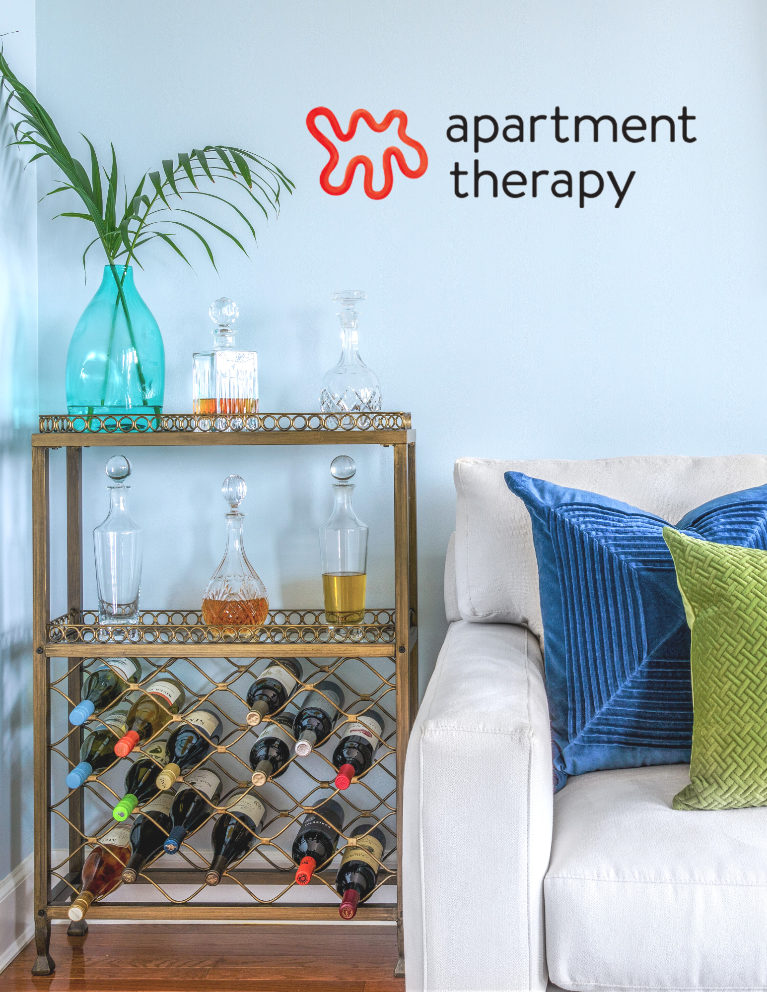 A gold metal wine rack with bottles of wine on a hardwood floor next to a white sofa with blue and green decorative pillows. A four-tiered glass decanter shelf holds glass decanters and glasses, topped with a turquoise vase and green palm leaves. A w