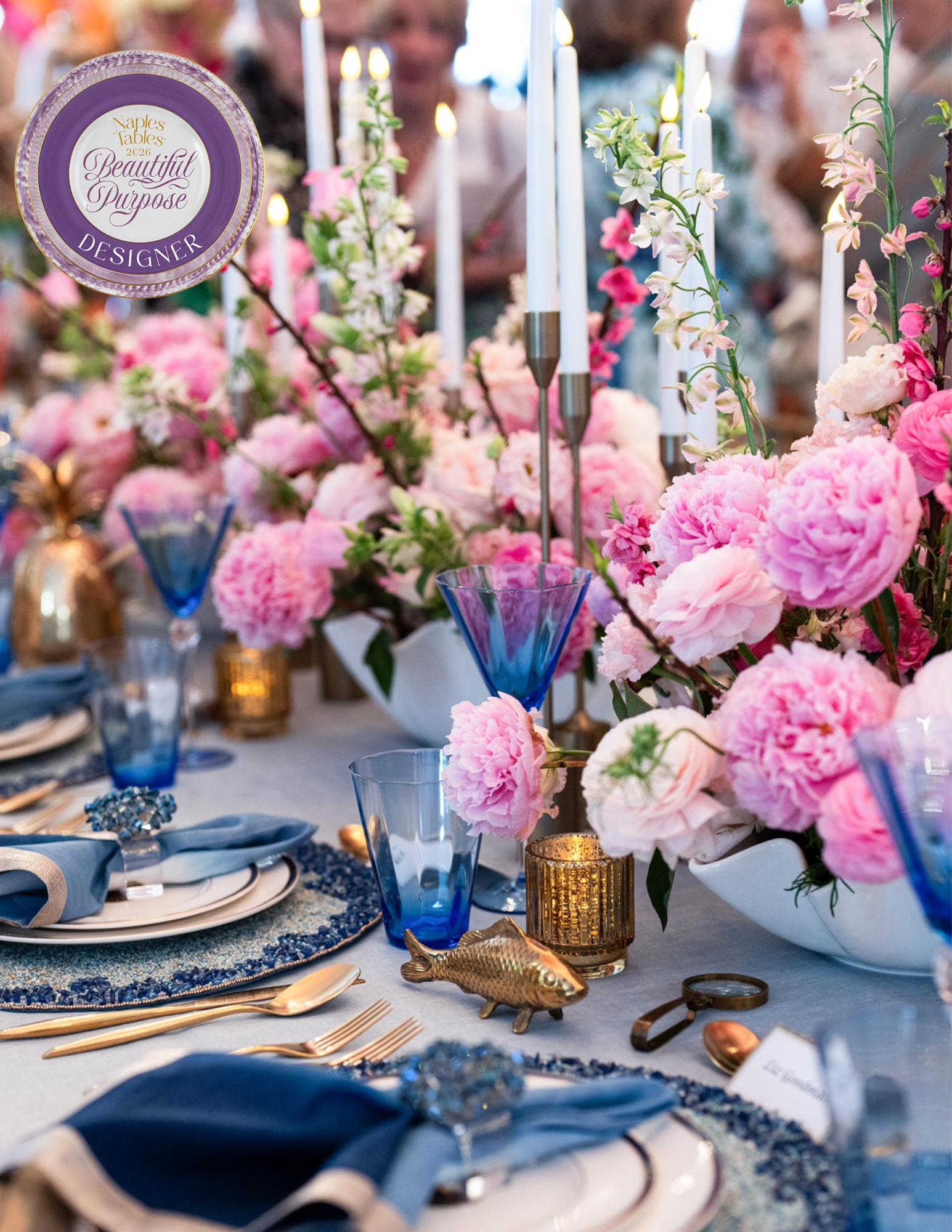 A decorated banquet table with pink floral arrangements, blue water glasses, gold utensils, and various candles, set for a formal event.
