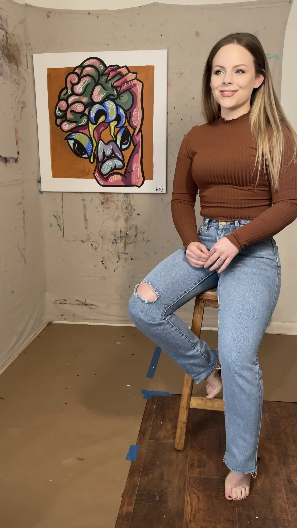 A woman sitting on a wooden stool next to a colorful abstract painting of a face on the wall.
