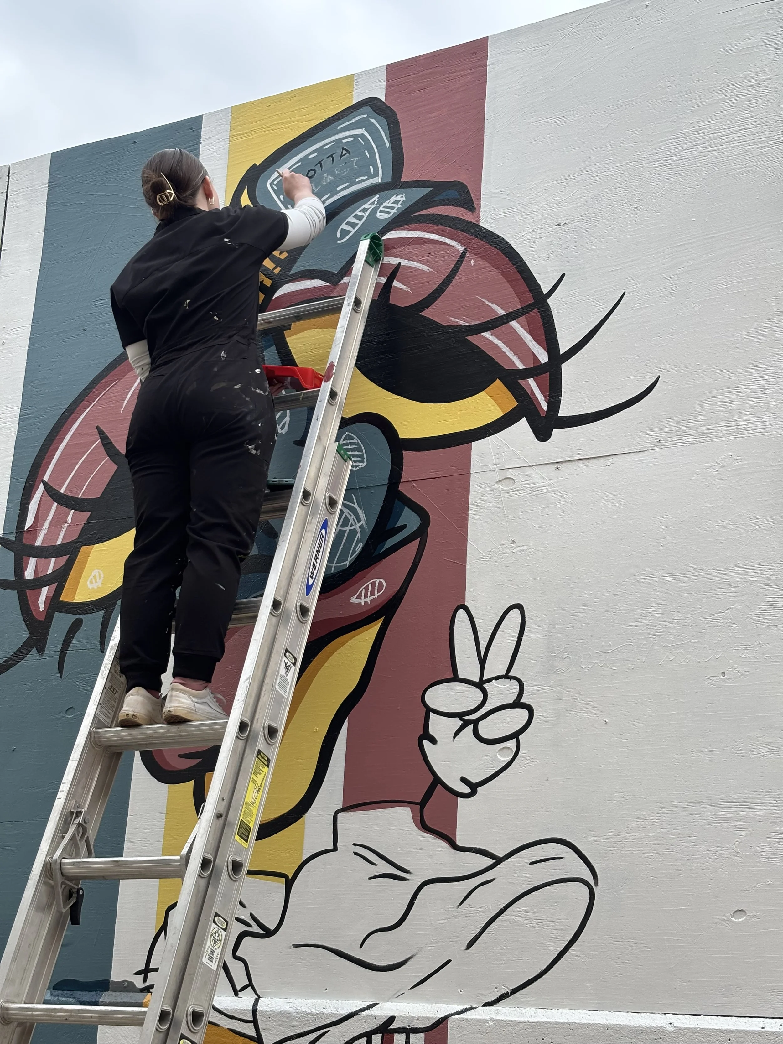 Artist painting a colorful mural on a large outdoor wall, depicting a cartoon character with sunglasses and a peace sign hand gesture.