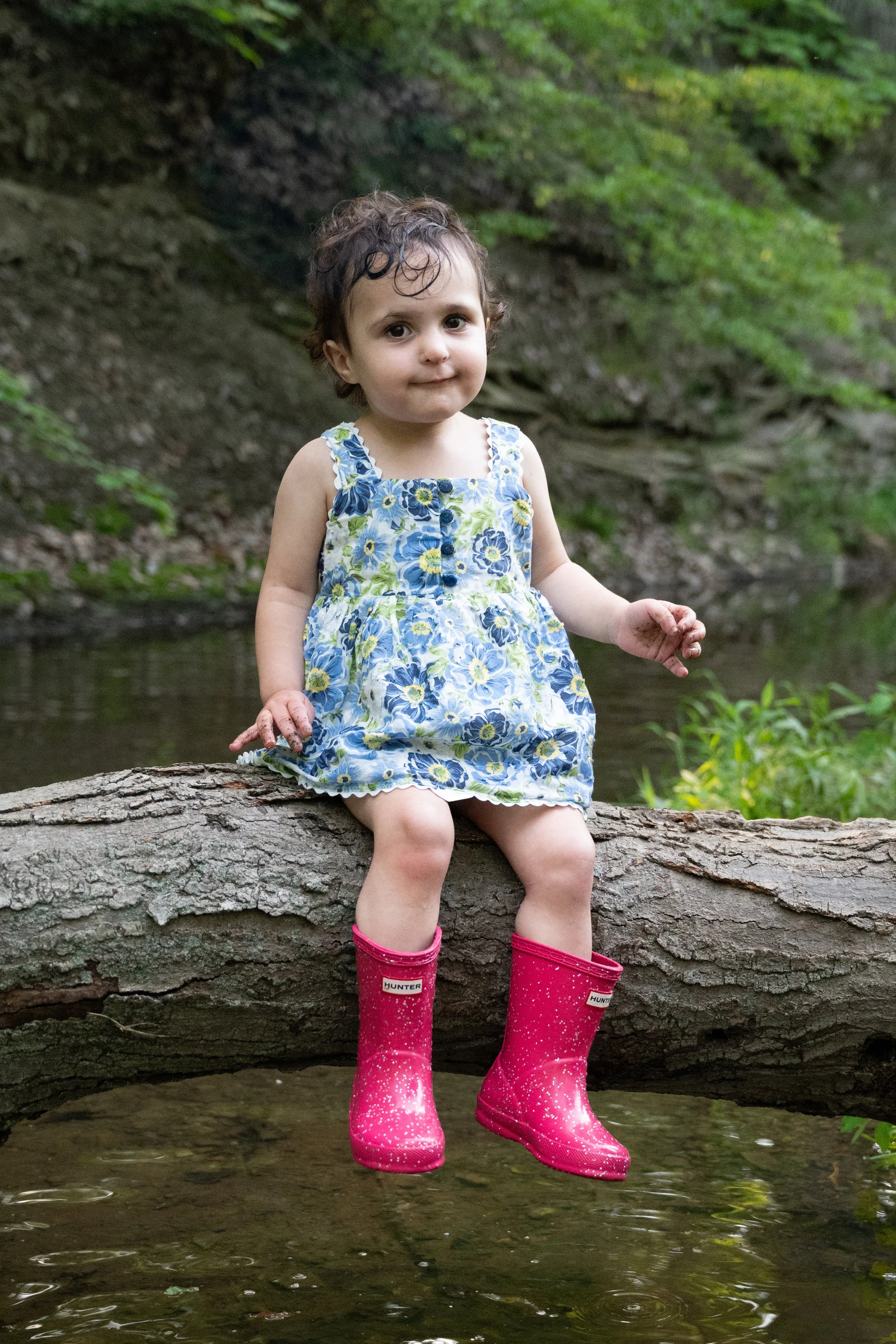 A young girl with curly hair sitting on a fallen tree trunk over a stream in a green forest, wearing a blue floral dress and bright pink rain boots.