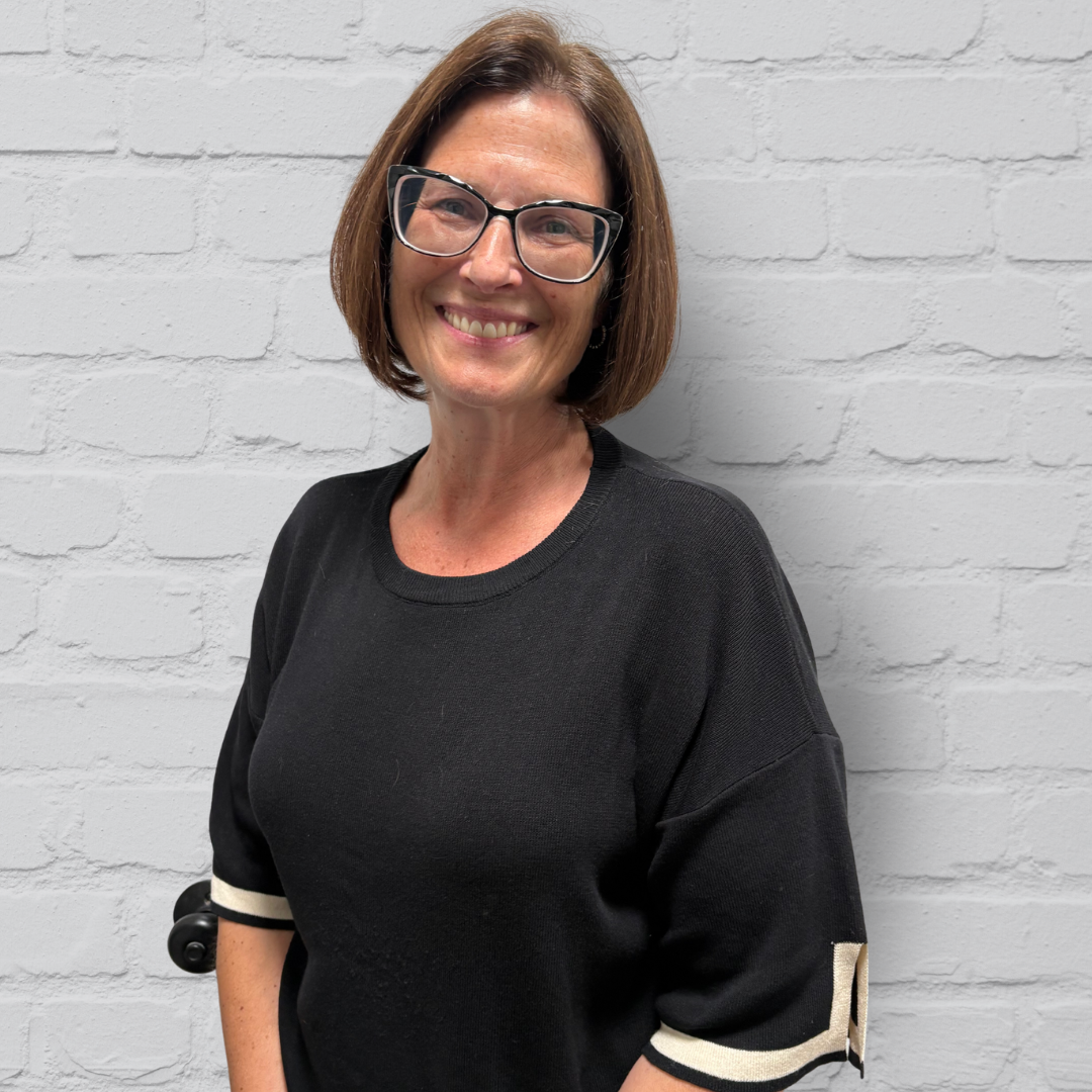 A woman with brown hair, glasses, and a black shirt smiling against a white brick wall.