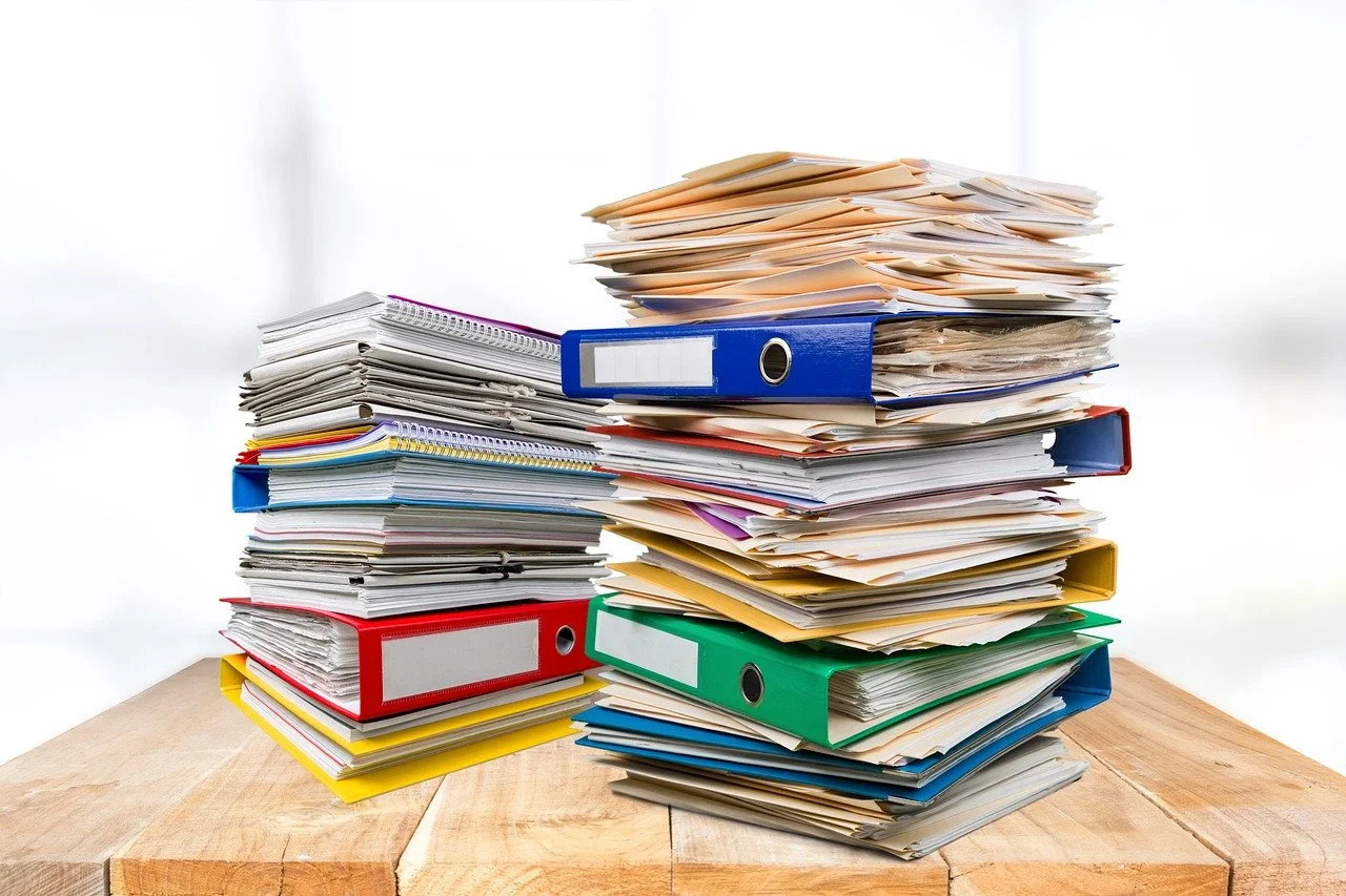 Multiple stacks of messy binders and papers on a wooden desk with a light background.