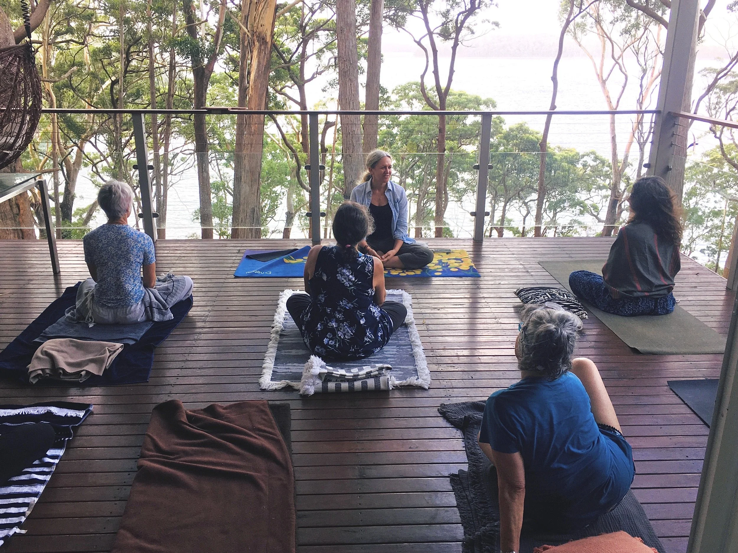 Allyson Lowbridge teaching a private group yoga class on a wooden deck overlooking the bush and the ocean.
